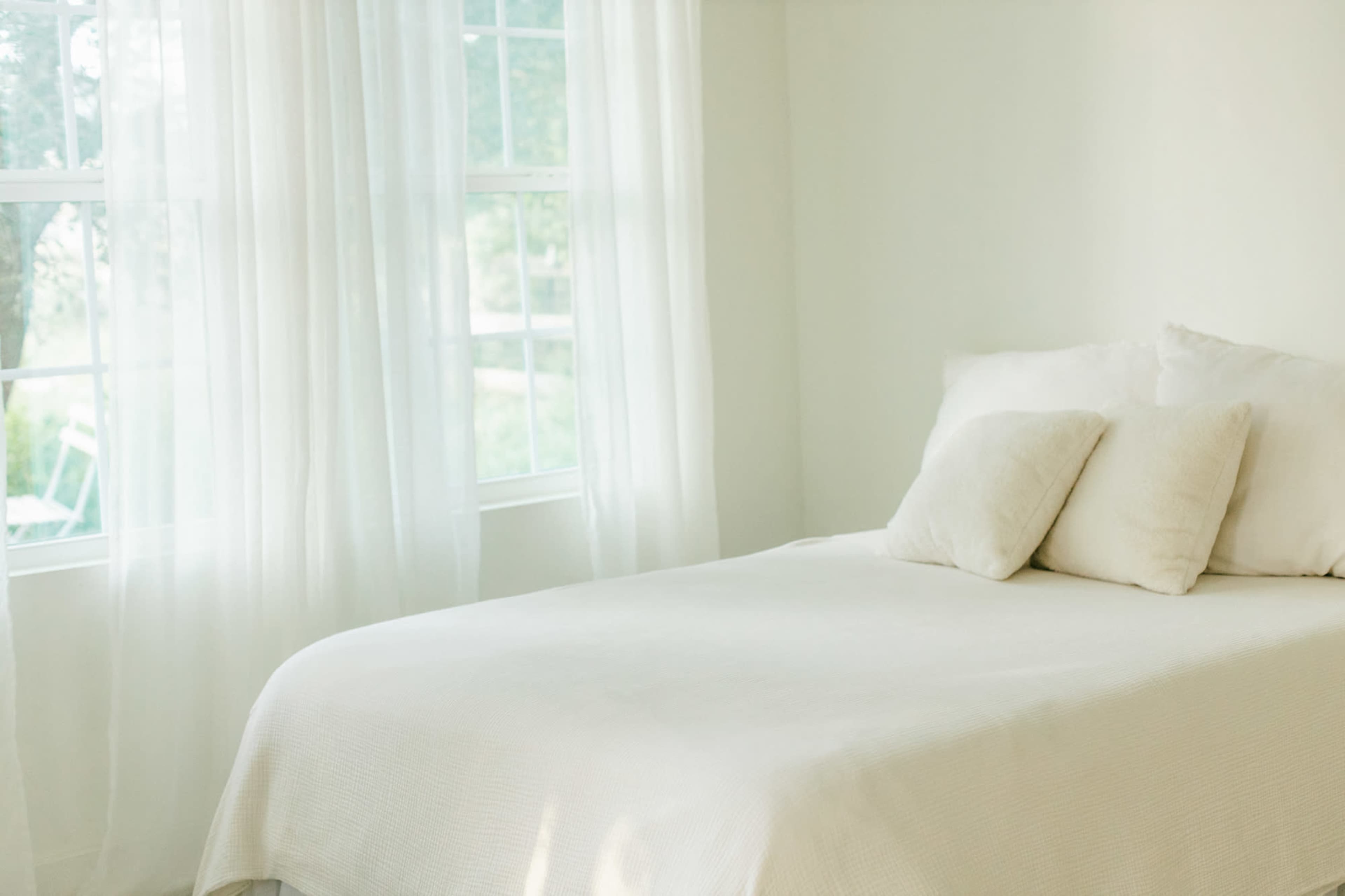 A neatly made bed with white bedding and two pillows is positioned next to a window with sheer white curtains.