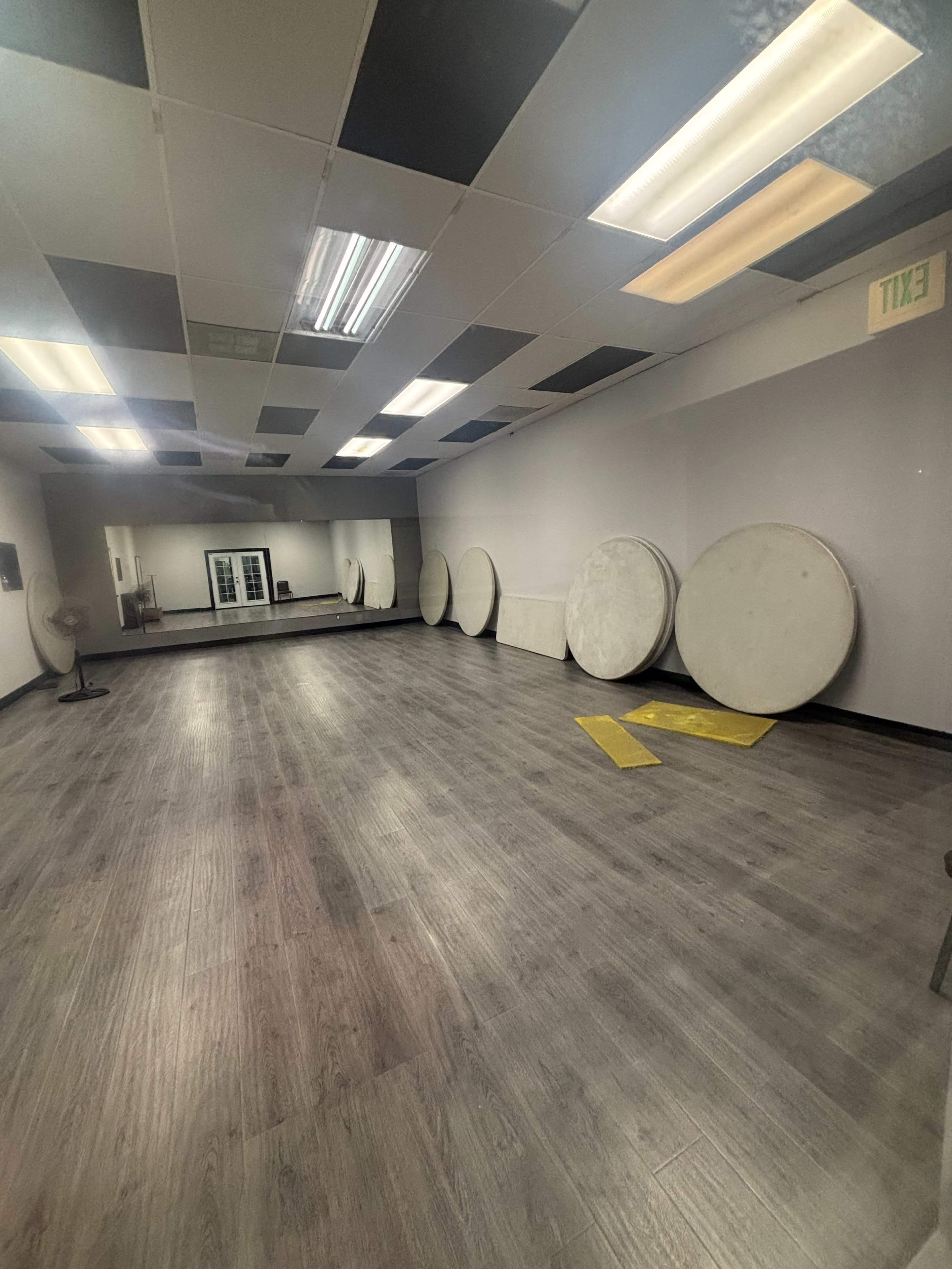 The image shows a spacious, empty room with a wooden floor and several large round tables leaning against the wall.