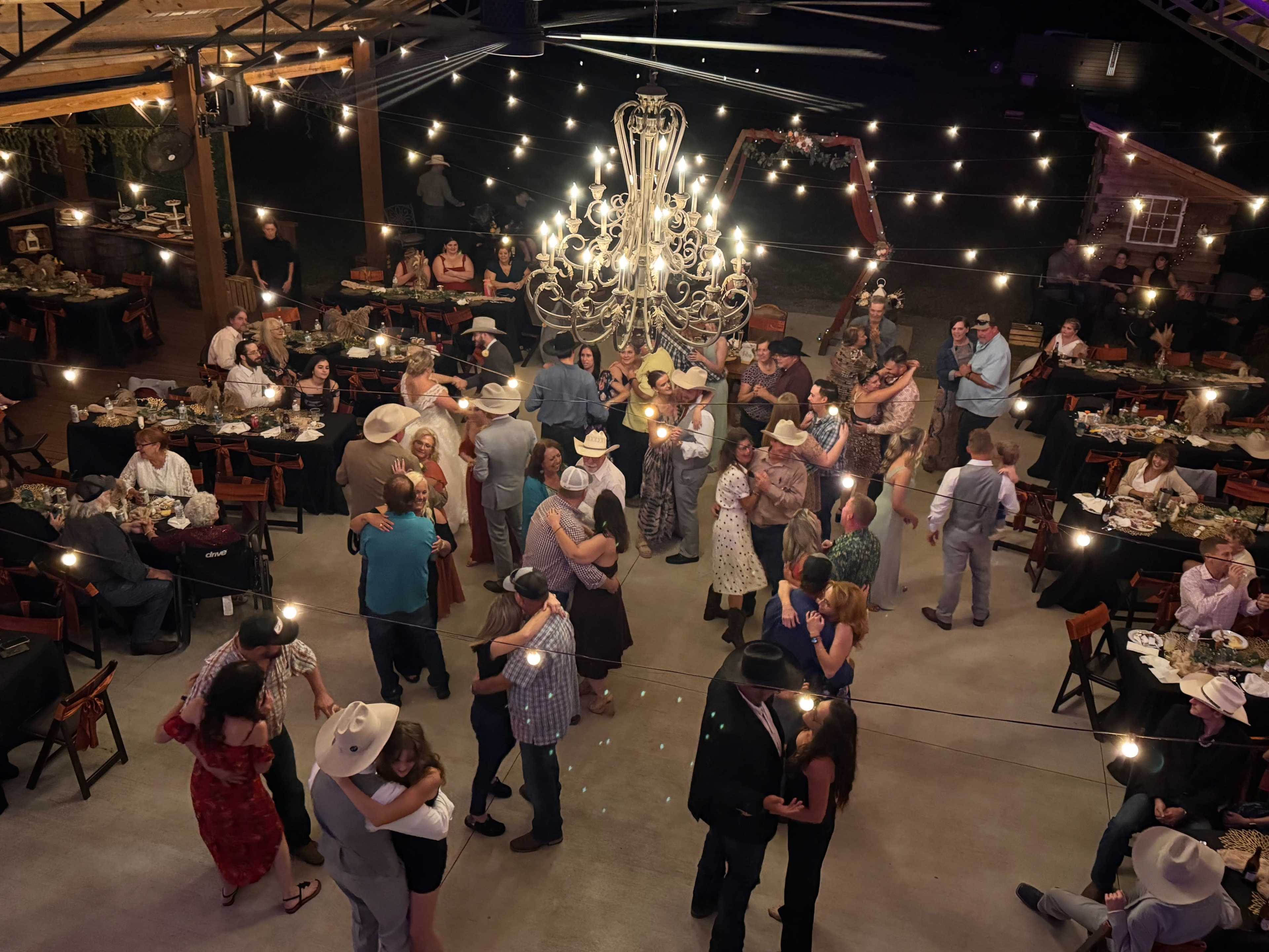 A large gathering of people wearing cowboy hats dances and mingles under string lights and a chandelier in a rustic venue with dining tables.