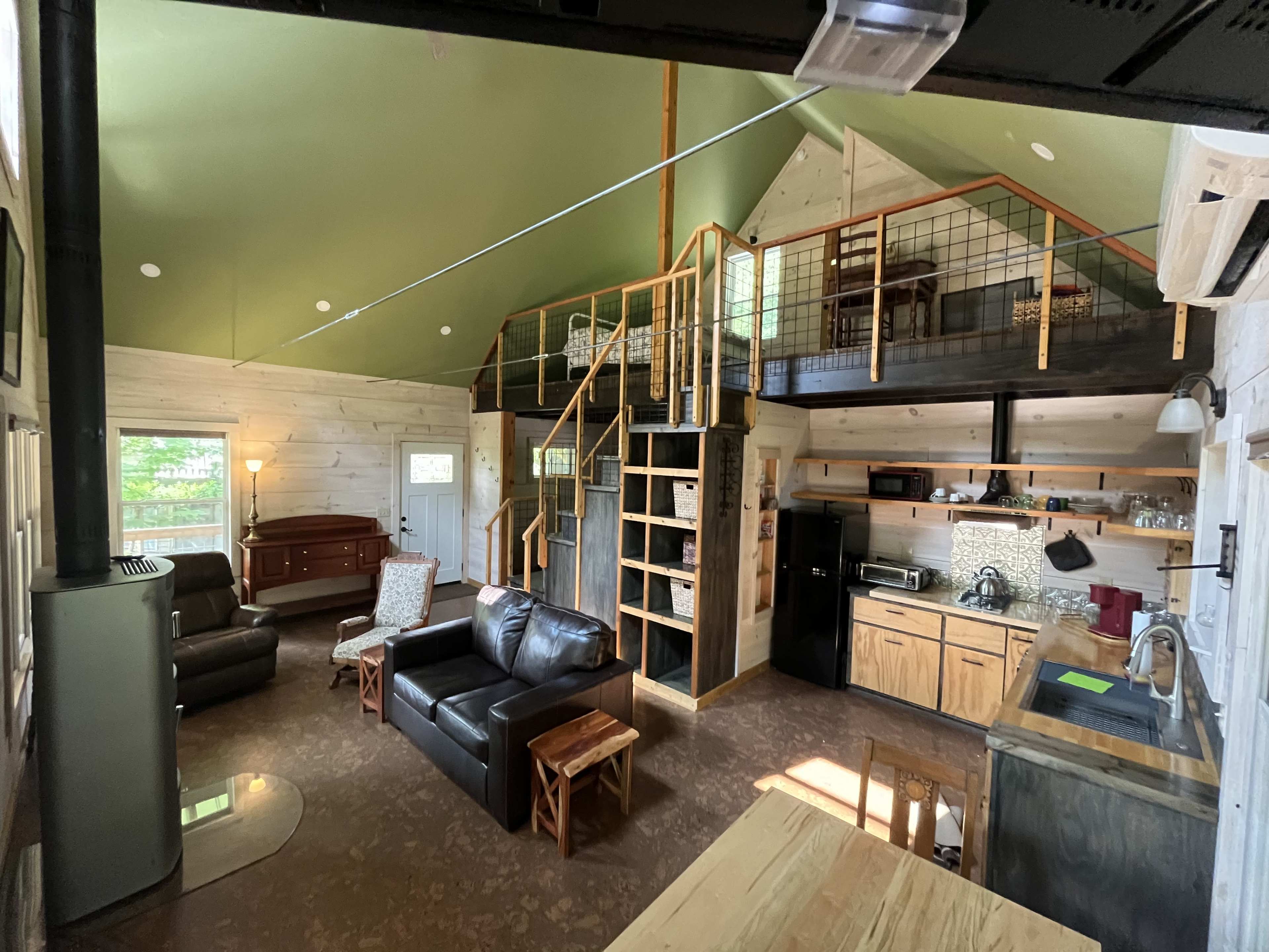 The image shows a modern cabin interior with a living area, kitchen, and a lofted sleeping space connected by a wooden staircase.