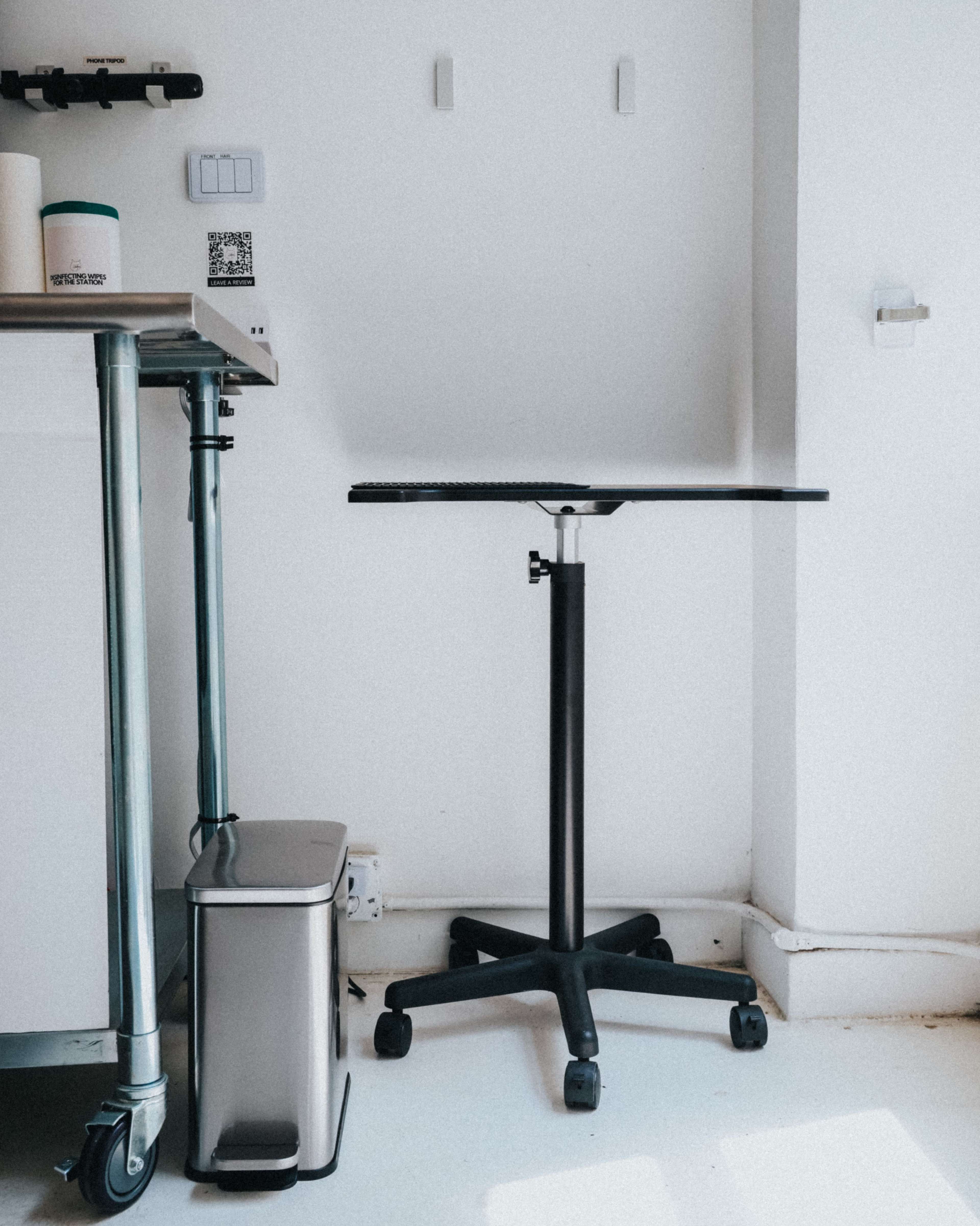 The image shows a minimalist workspace featuring a black height-adjustable table on a wheeled base, a metal trash bin, and a countertop with a QR code scanner.