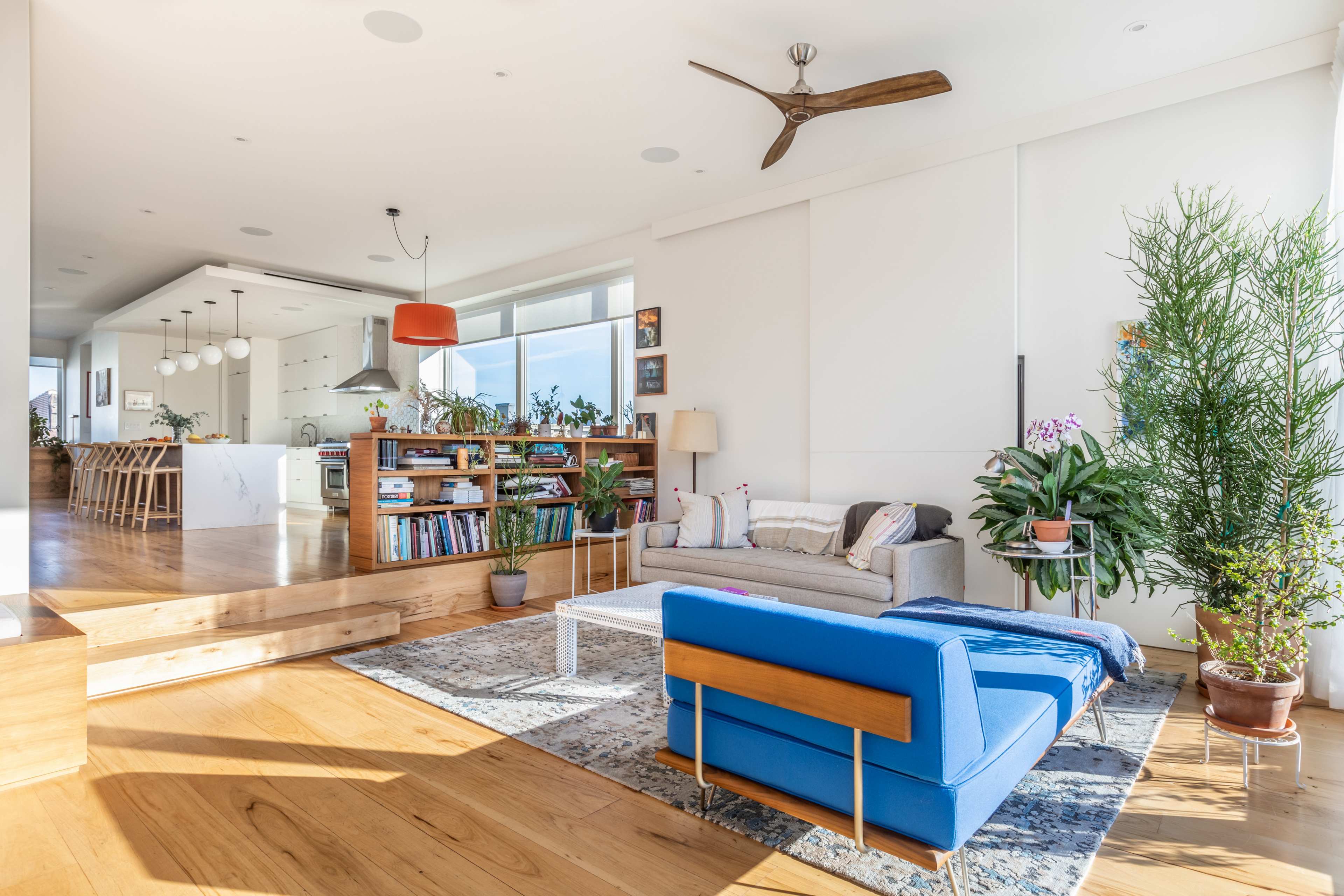 A bright and modern living room featuring a blue sofa, a coffee table, and a bookshelf, with an open kitchen visible in the background.