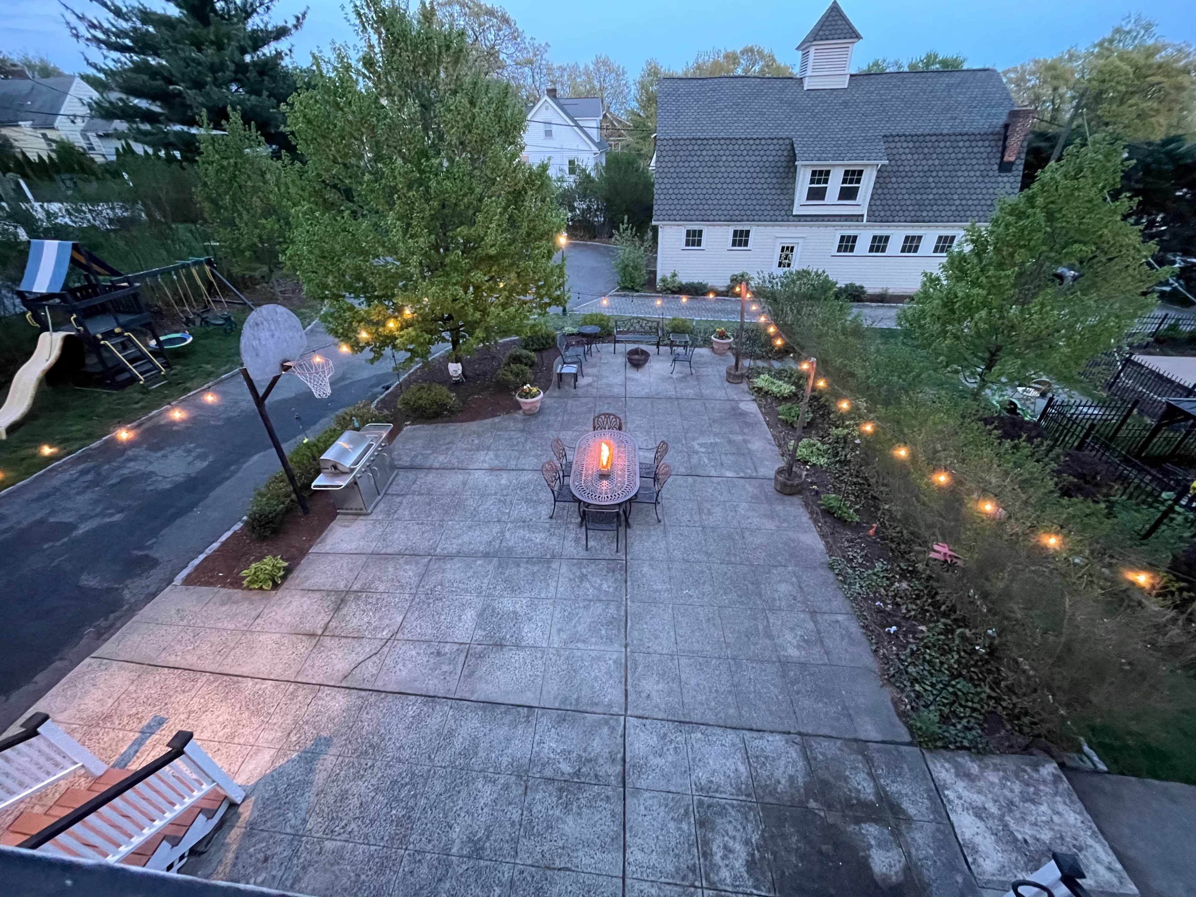 The image shows a spacious backyard patio area with a central table and chairs, surrounded by trees and outdoor lights, leading to a house in the background.