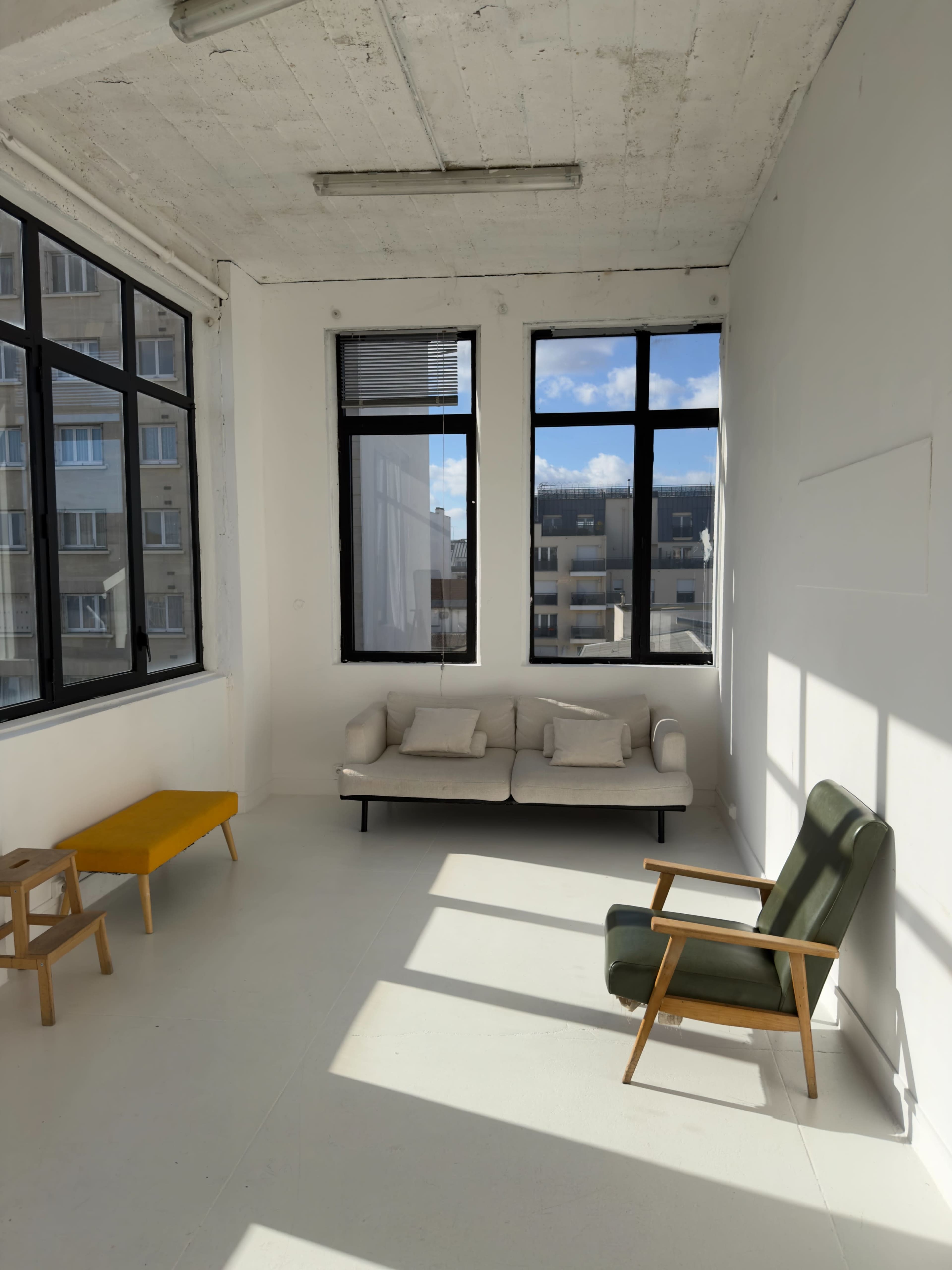 The image shows a minimalist interior with a white wall, large windows, a light-colored sofa, a green armchair, and a small yellow side table.