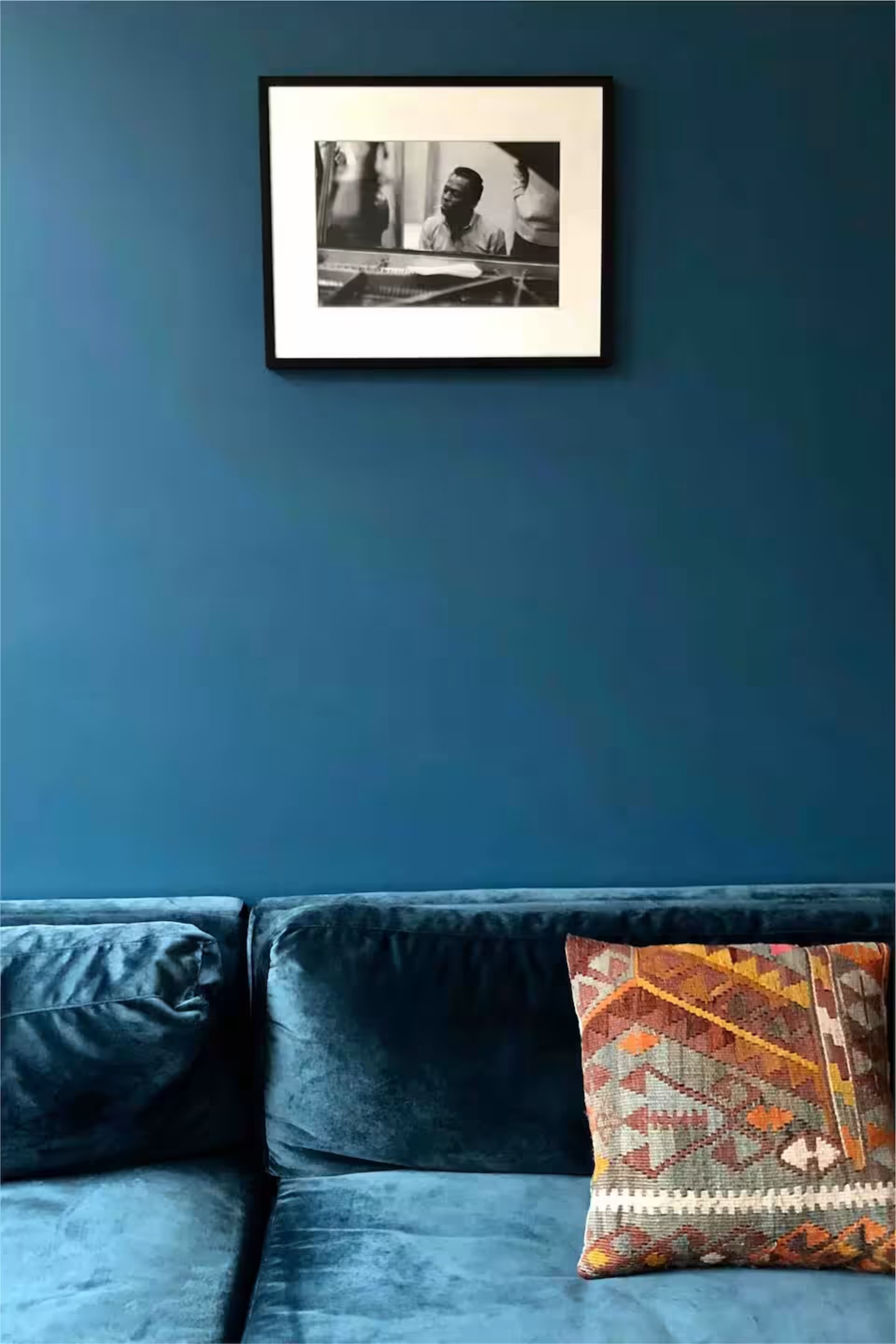 The image shows a blue wall featuring a framed black-and-white photograph above a plush teal couch adorned with a patterned throw pillow.