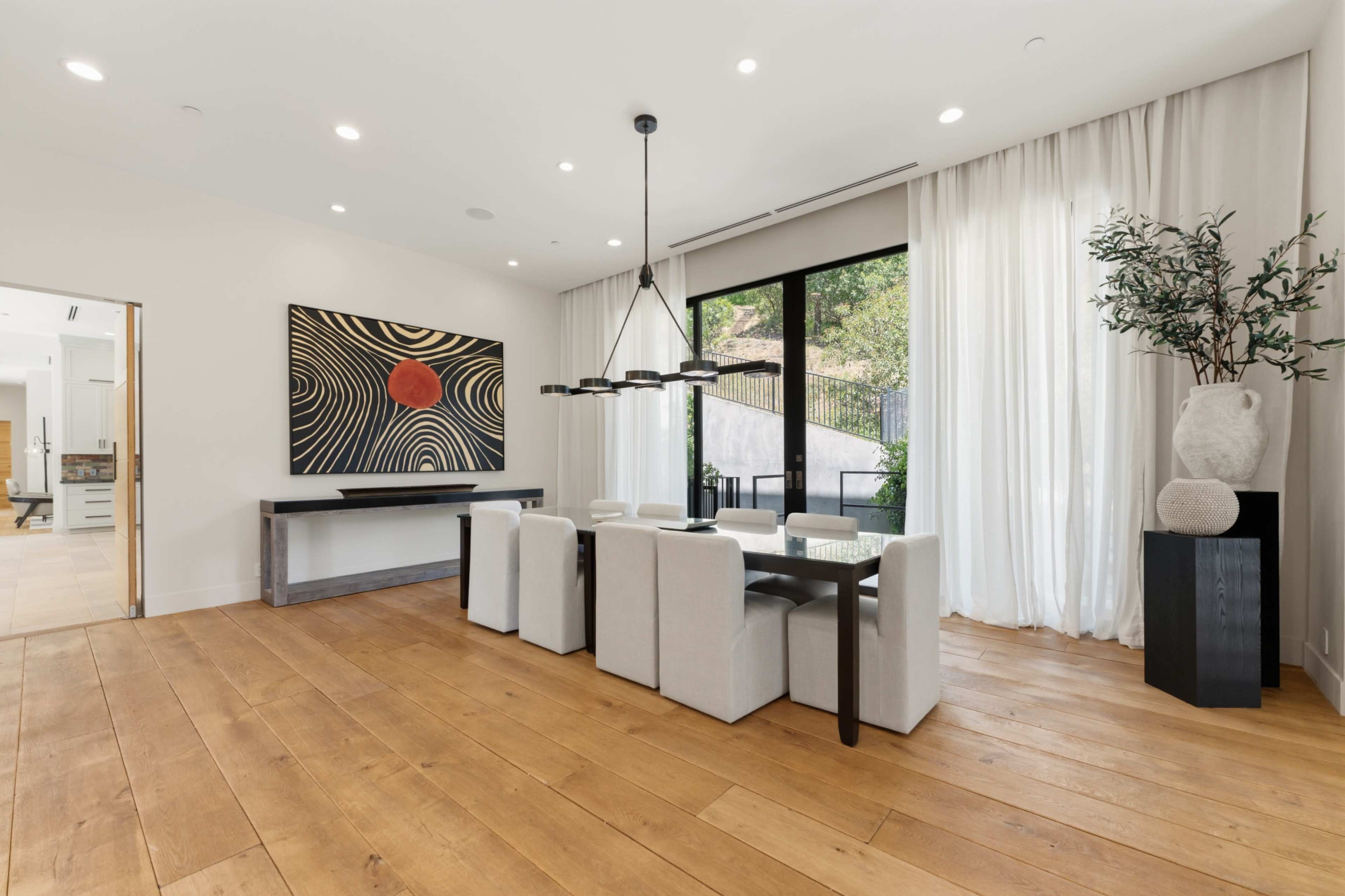 A modern dining area features a rectangular table surrounded by six light-colored chairs, with large windows showcasing an outdoor view and a bold piece of abstract art on the wall.