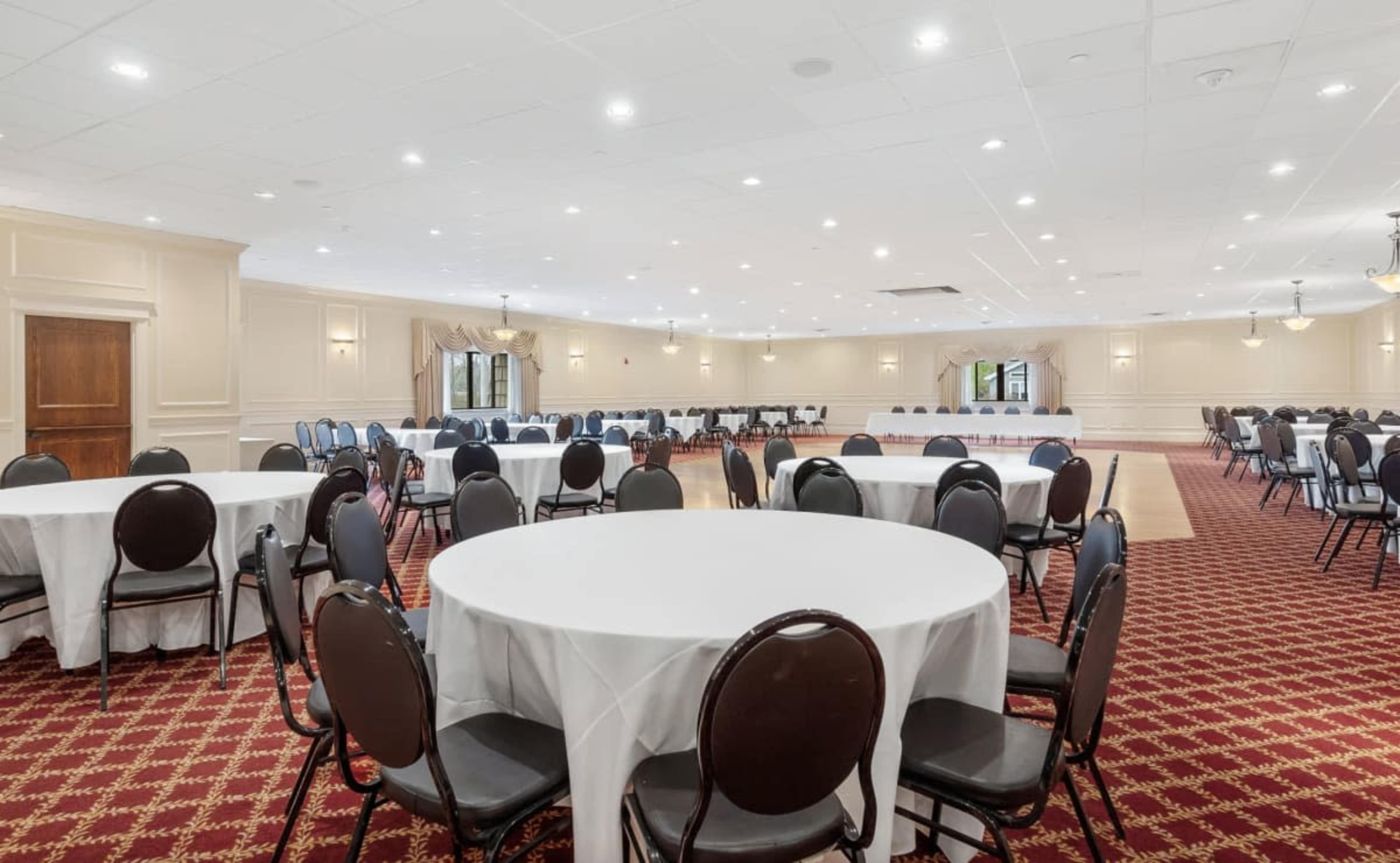 The image shows a large event room set up with round tables covered in white tablecloths and surrounded by black chairs, with an open space in the center.