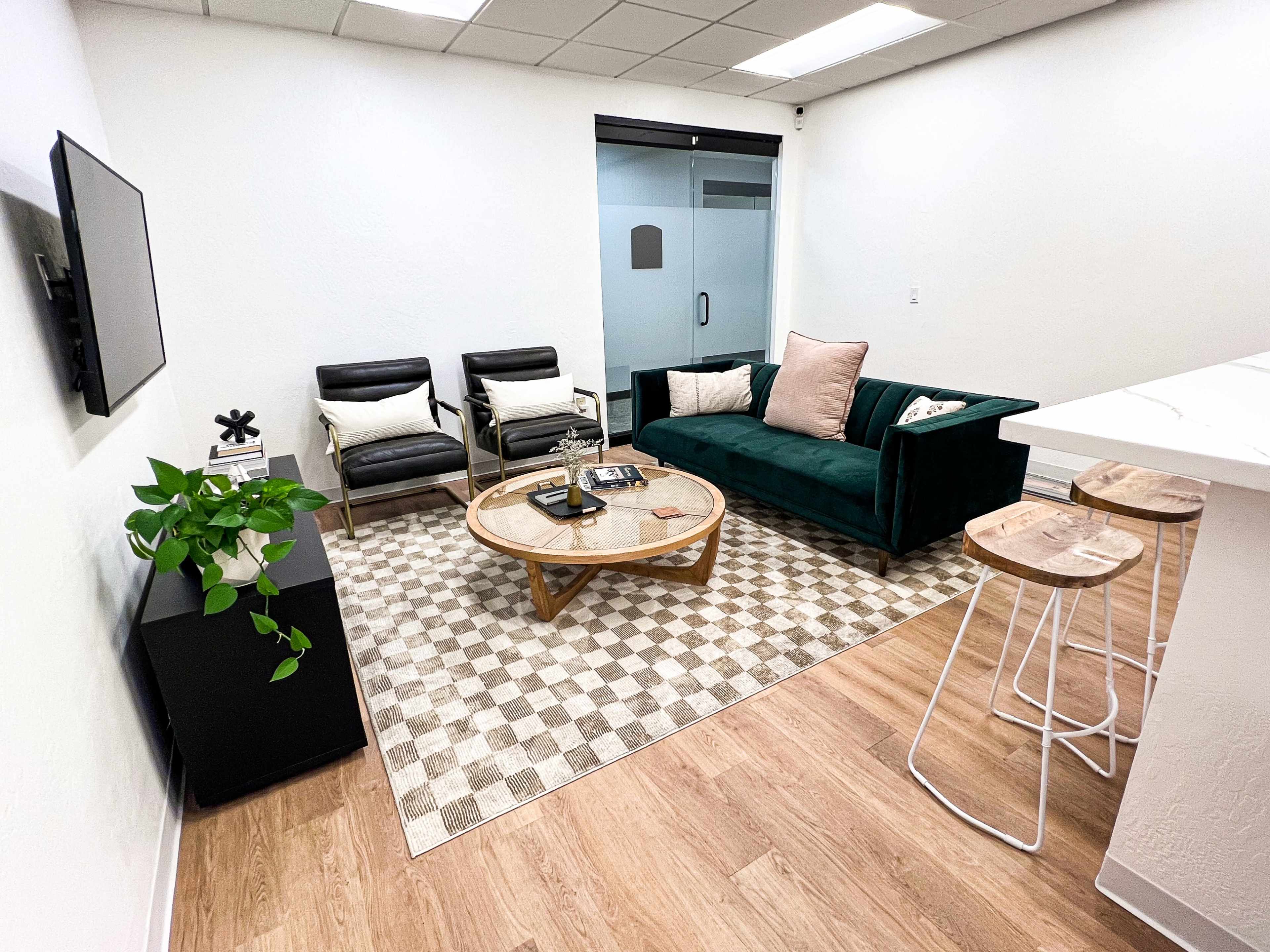 A modern, minimalistic lounge area with two black chairs, a green sofa, a round coffee table on a patterned rug, and a small plant next to a black cabinet.
