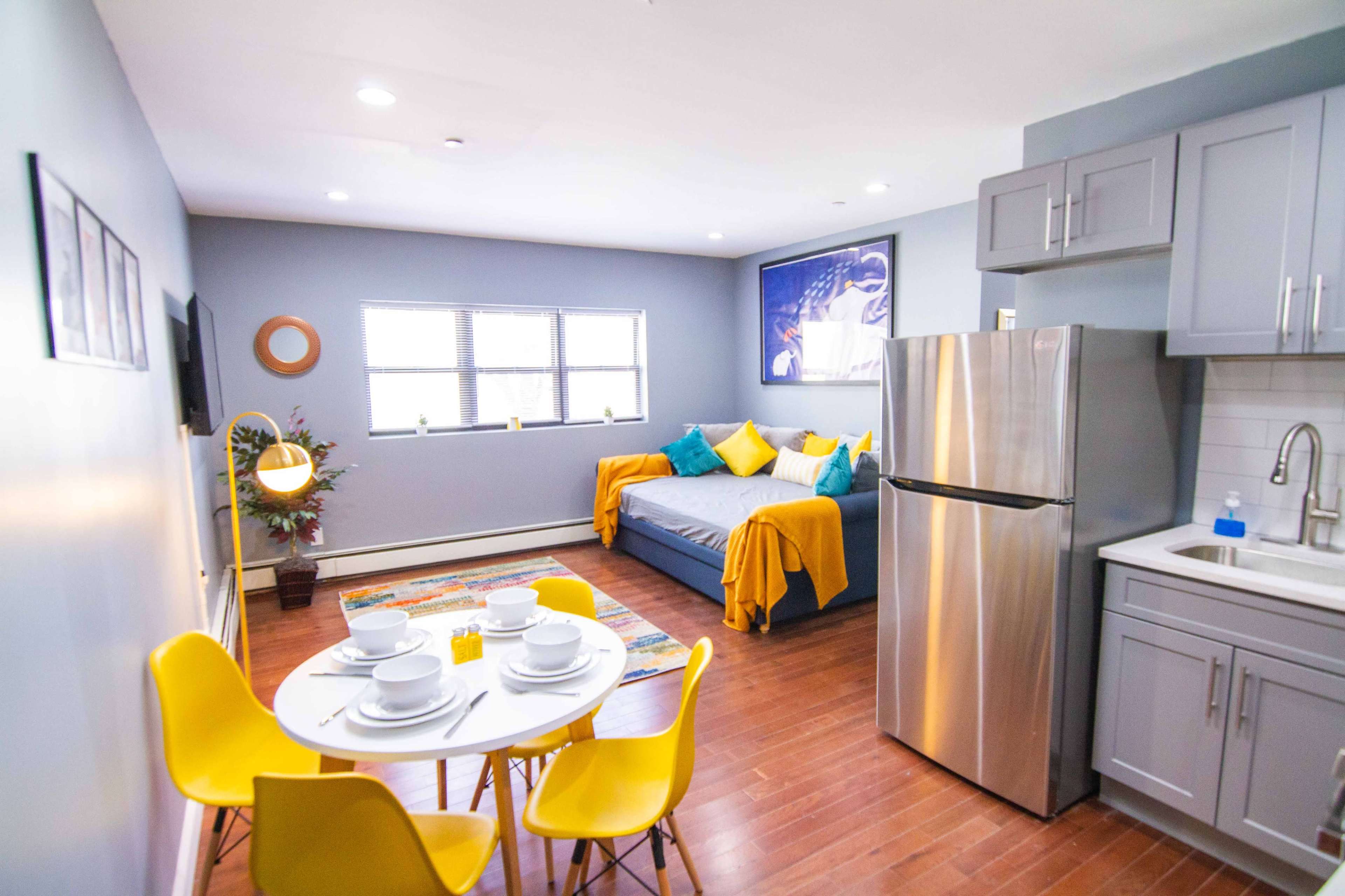 A modern small living space featuring a kitchen area with gray cabinets, a stainless steel refrigerator, a circular dining table with yellow chairs, and a seating area with a blue couch adorned with colorful pillows.