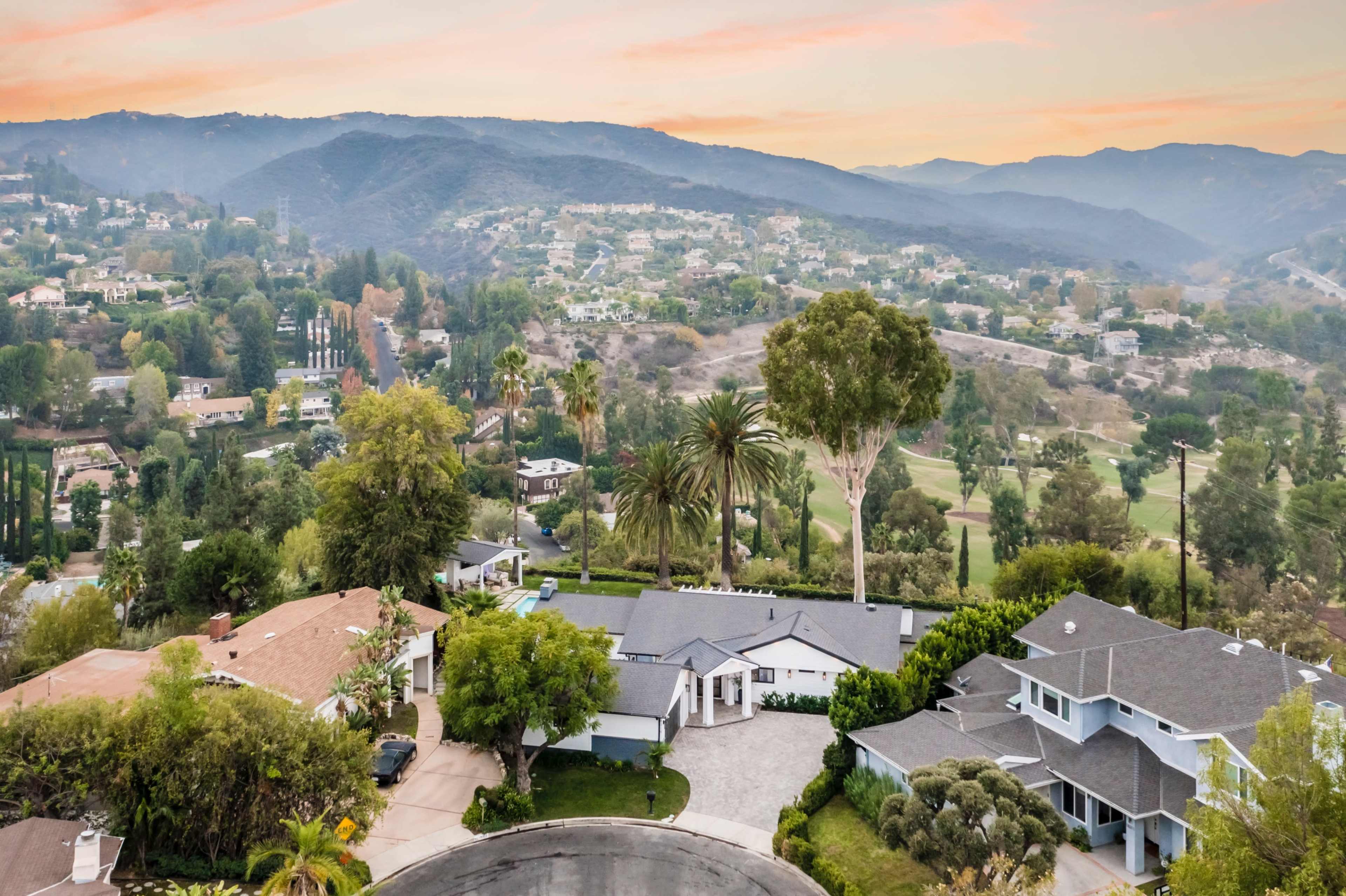 A residential area with several houses, surrounded by greenery and hills in the background.