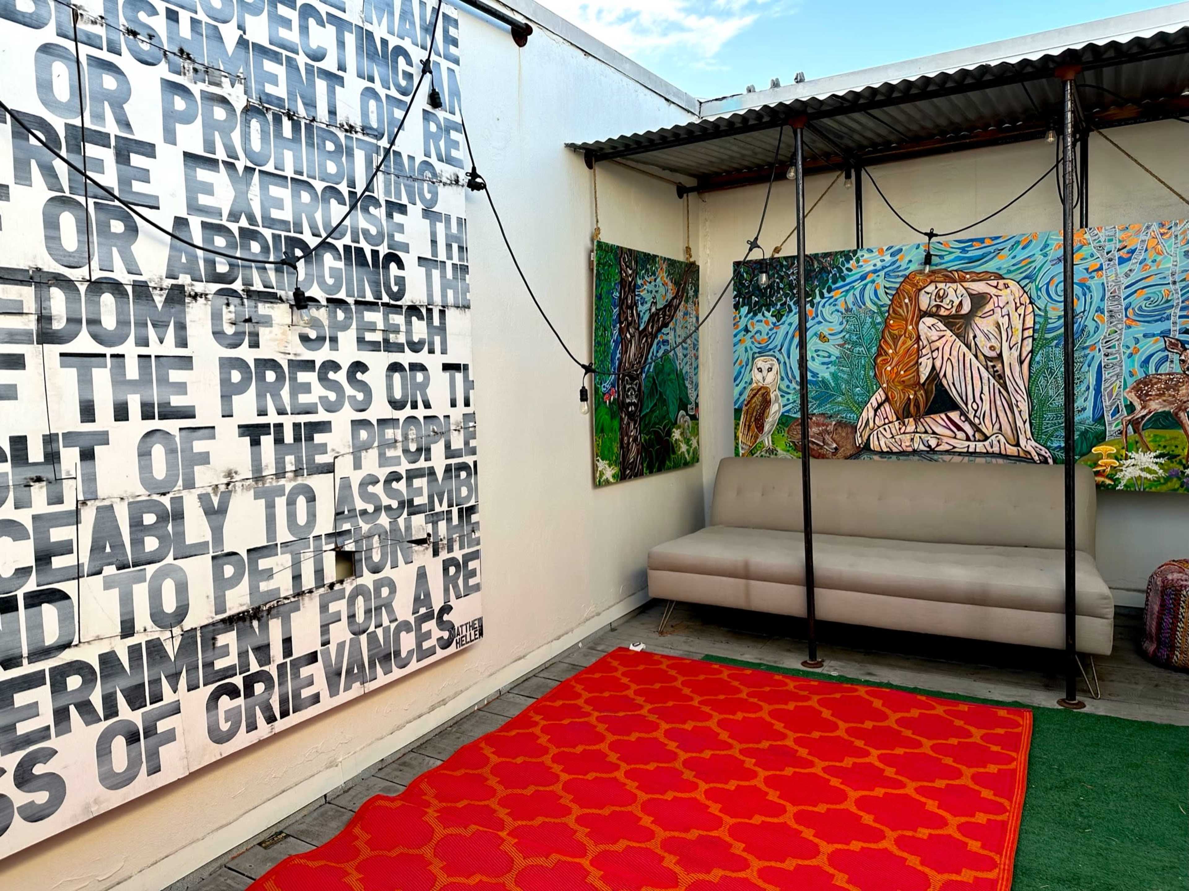 A rooftop space with a large wall displaying text, colorful artwork on the adjacent walls, a gray sofa, and a patterned red rug on the floor.
