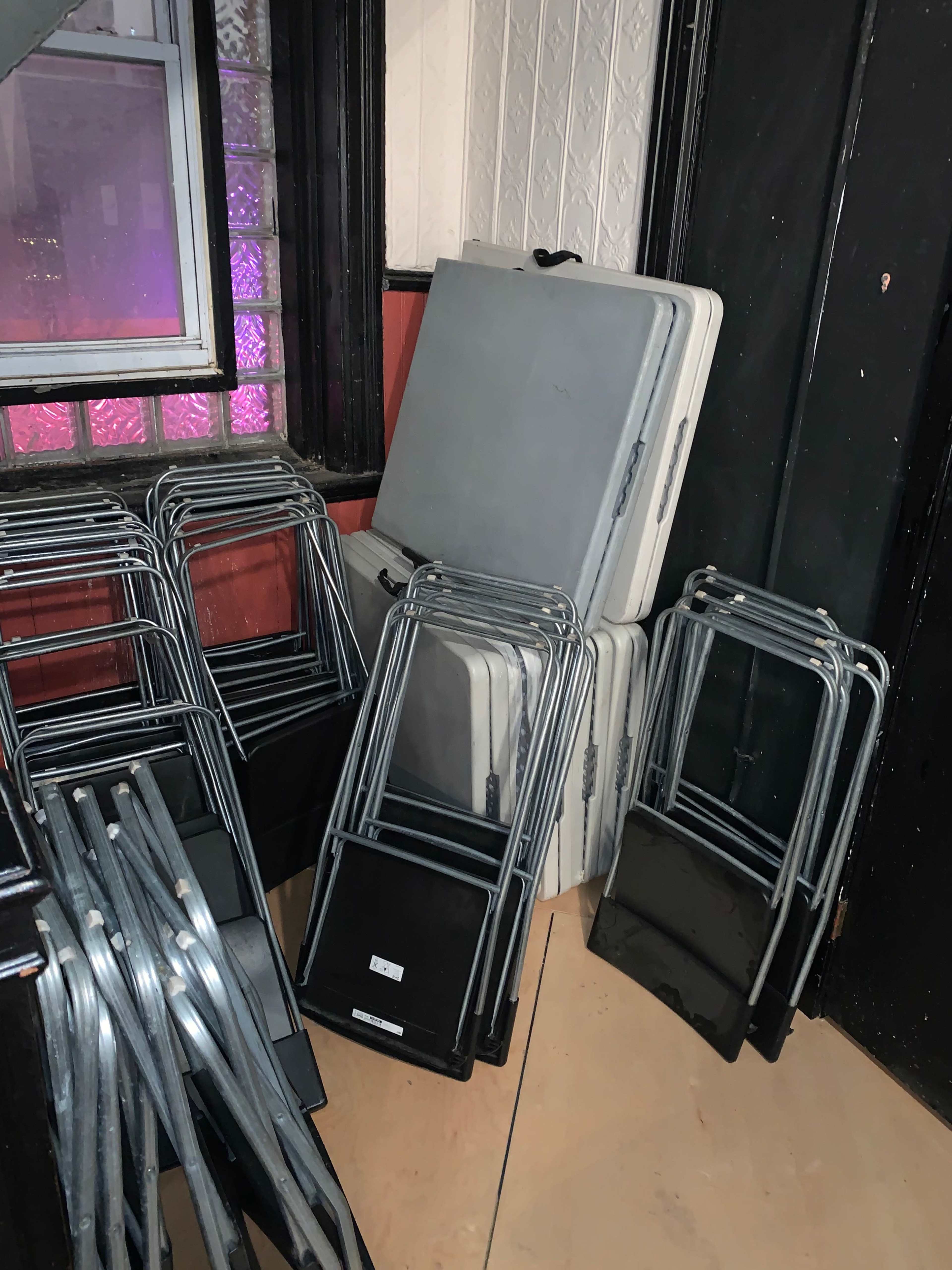 A stack of folded tables and several metal-framed chairs are arranged against a wall in a dimly lit room.