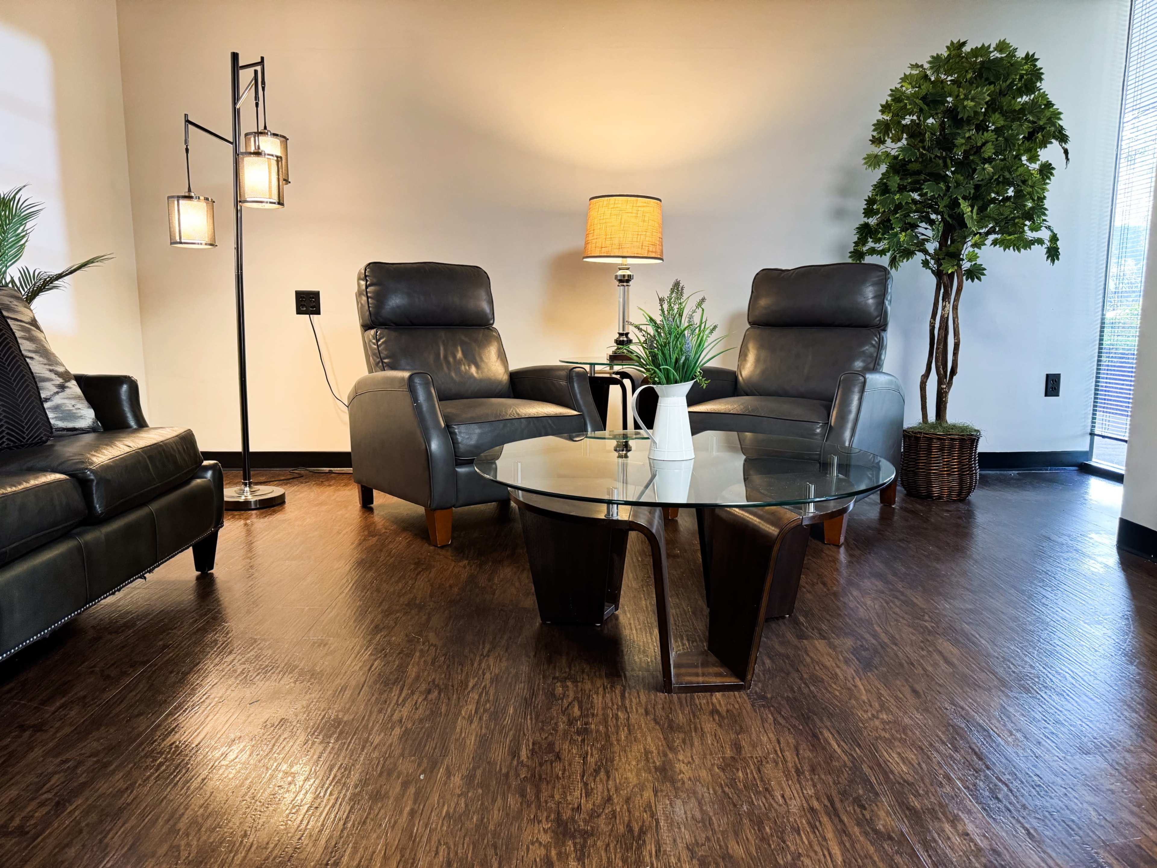 The image shows a modern lounge area featuring two gray armchairs, a round glass table, a lamp, and a potted plant.