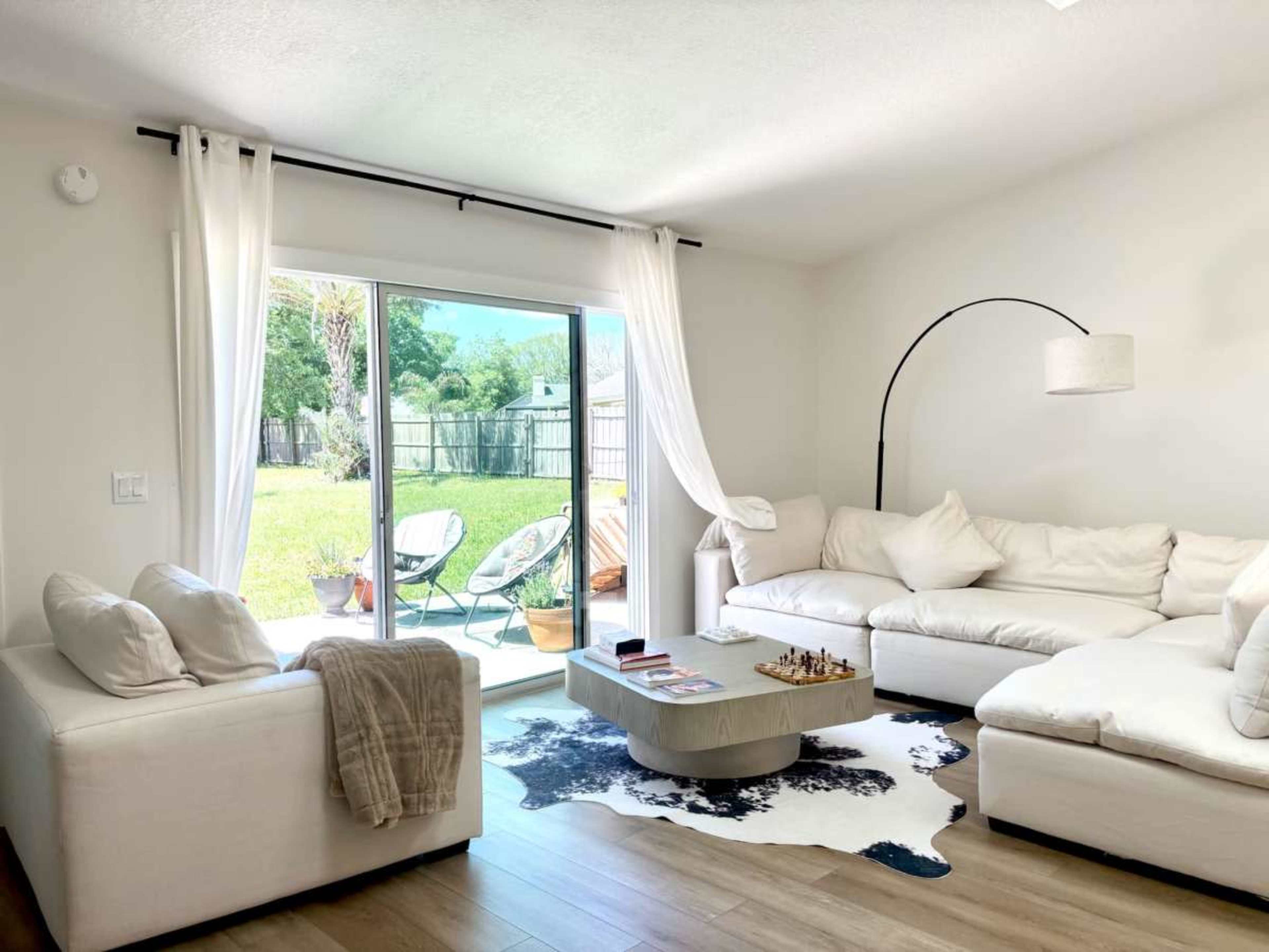 The image shows a bright living room with a white sectional sofa, a coffee table on a cowhide rug, and sliding glass doors leading to a grassy backyard.