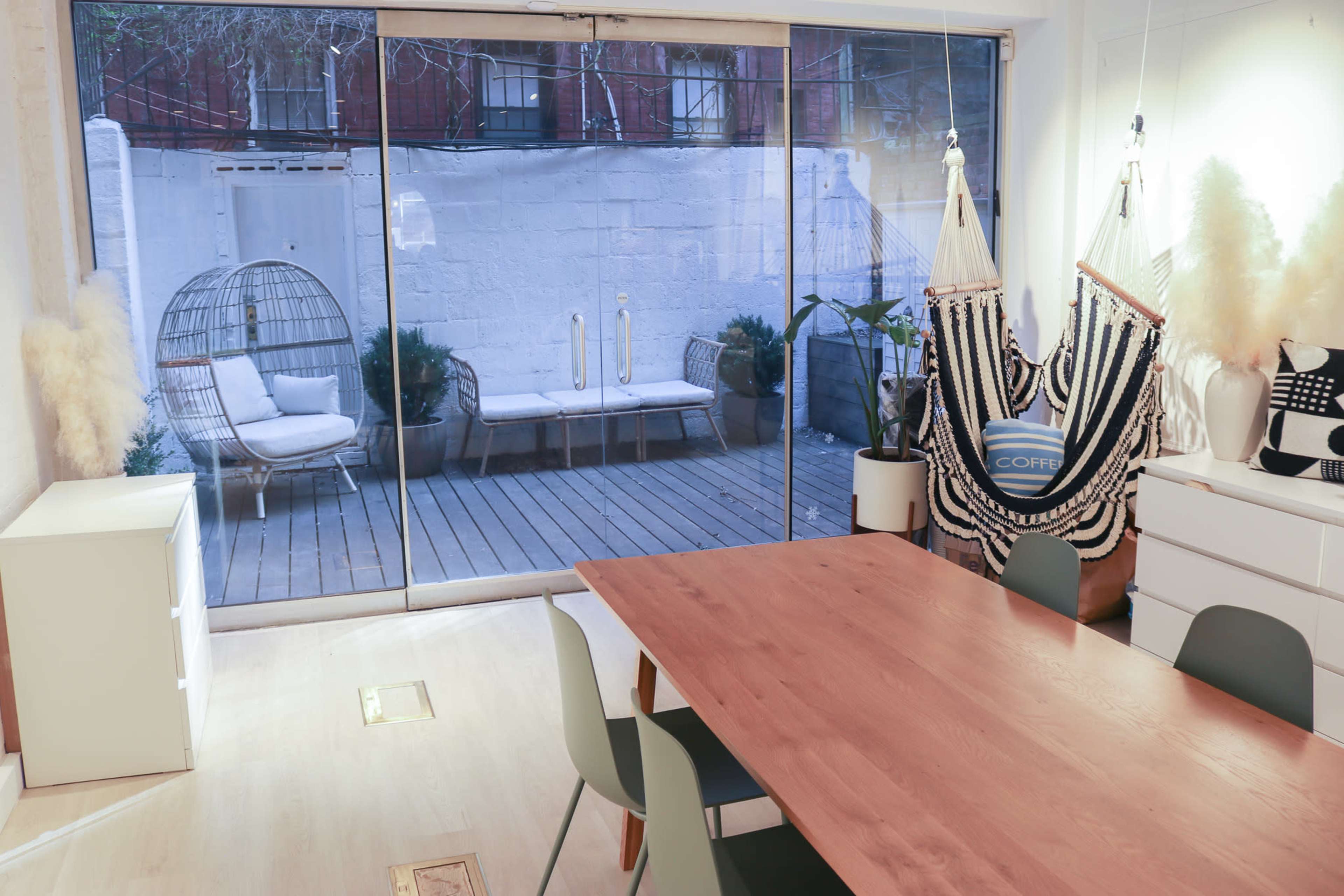 A modern interior room features a wooden table surrounded by green chairs, with a large glass door that opens to a wooden deck and outdoor seating area.