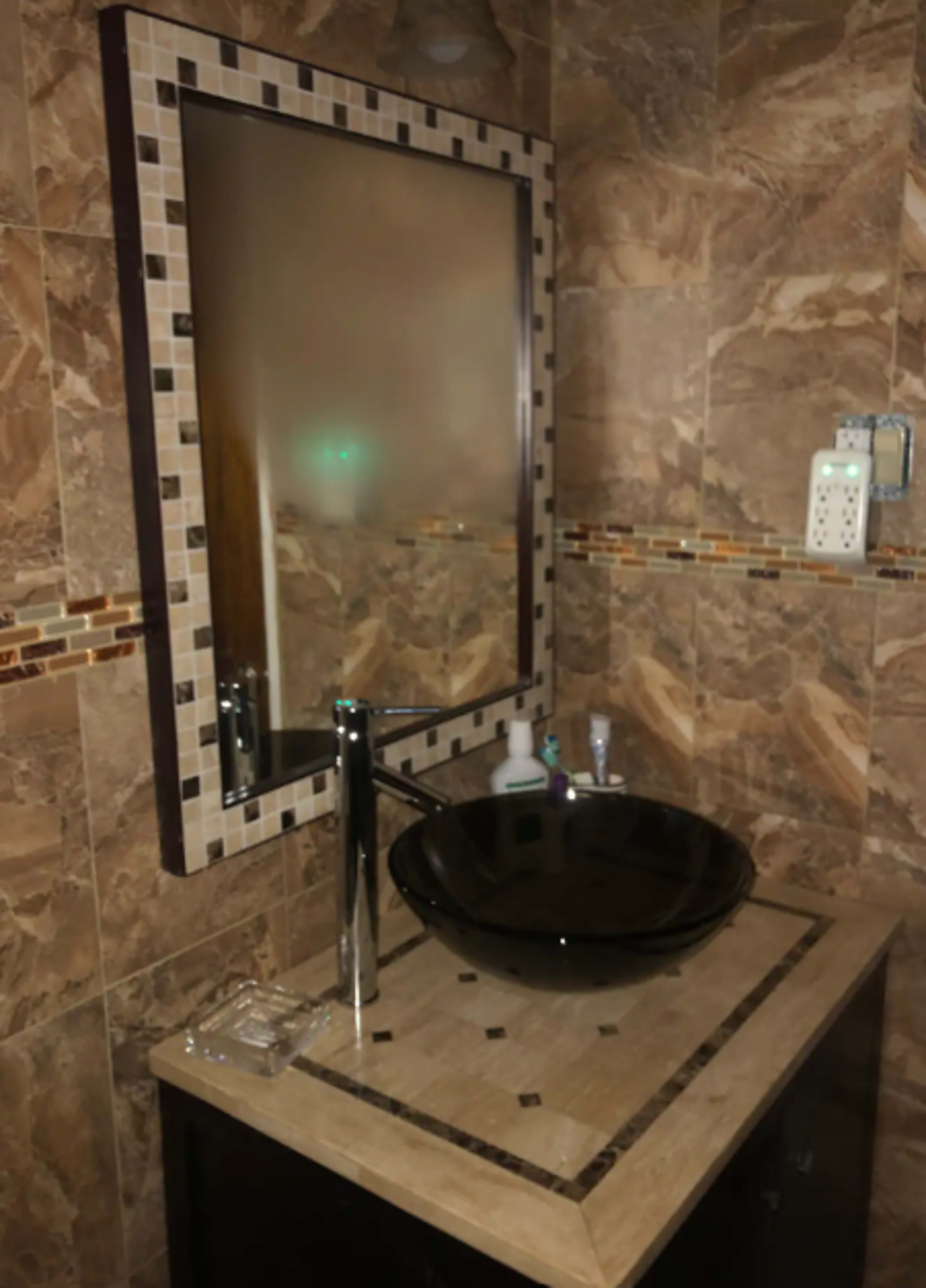 A modern bathroom with a black vessel sink, a rectangular mirror framed with decorative tiles, and marble-patterned walls.