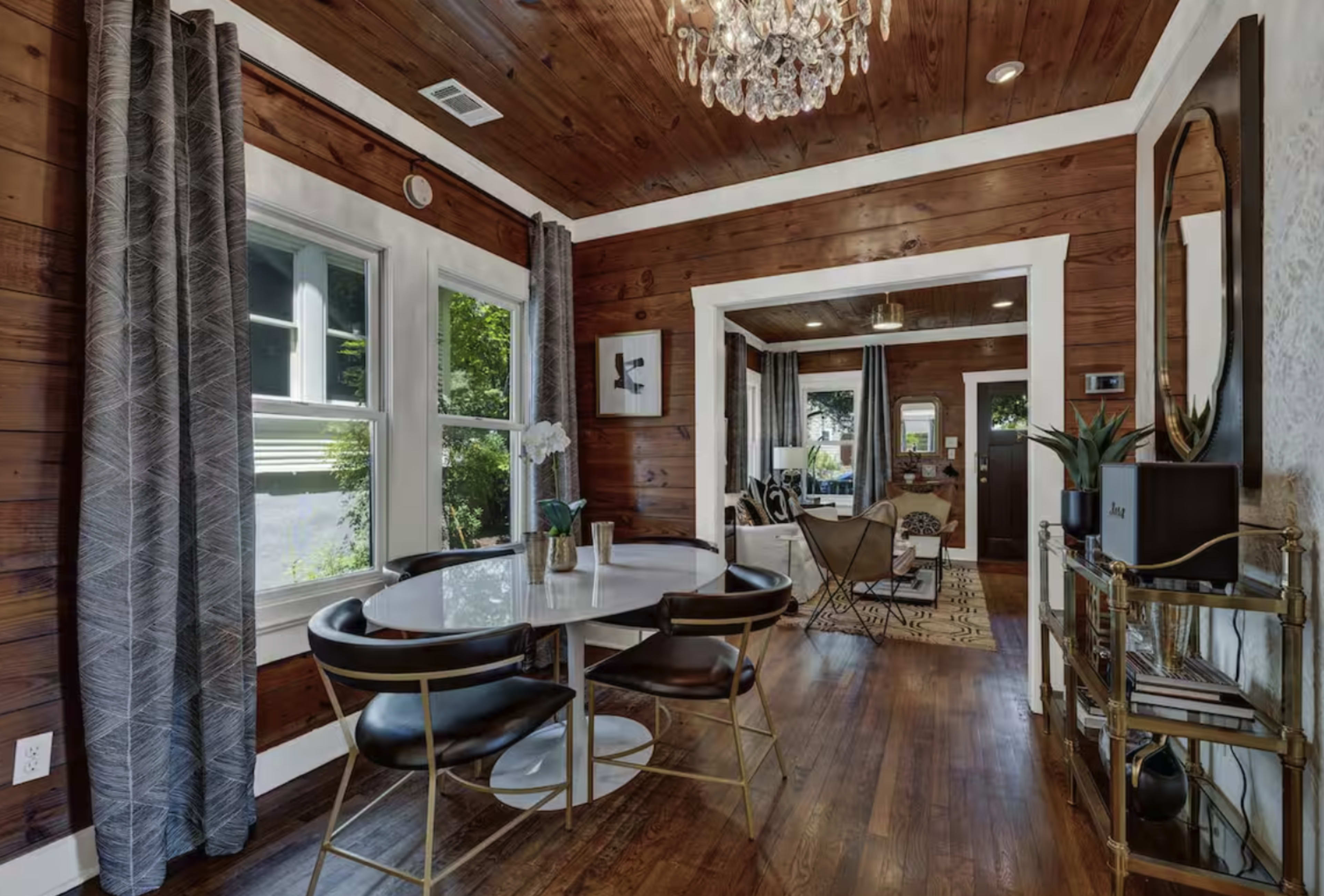 The image shows a dining area with a round table and black chairs, featuring wooden walls and a chandelier, opening into a living space decorated with plants and contemporary furniture.