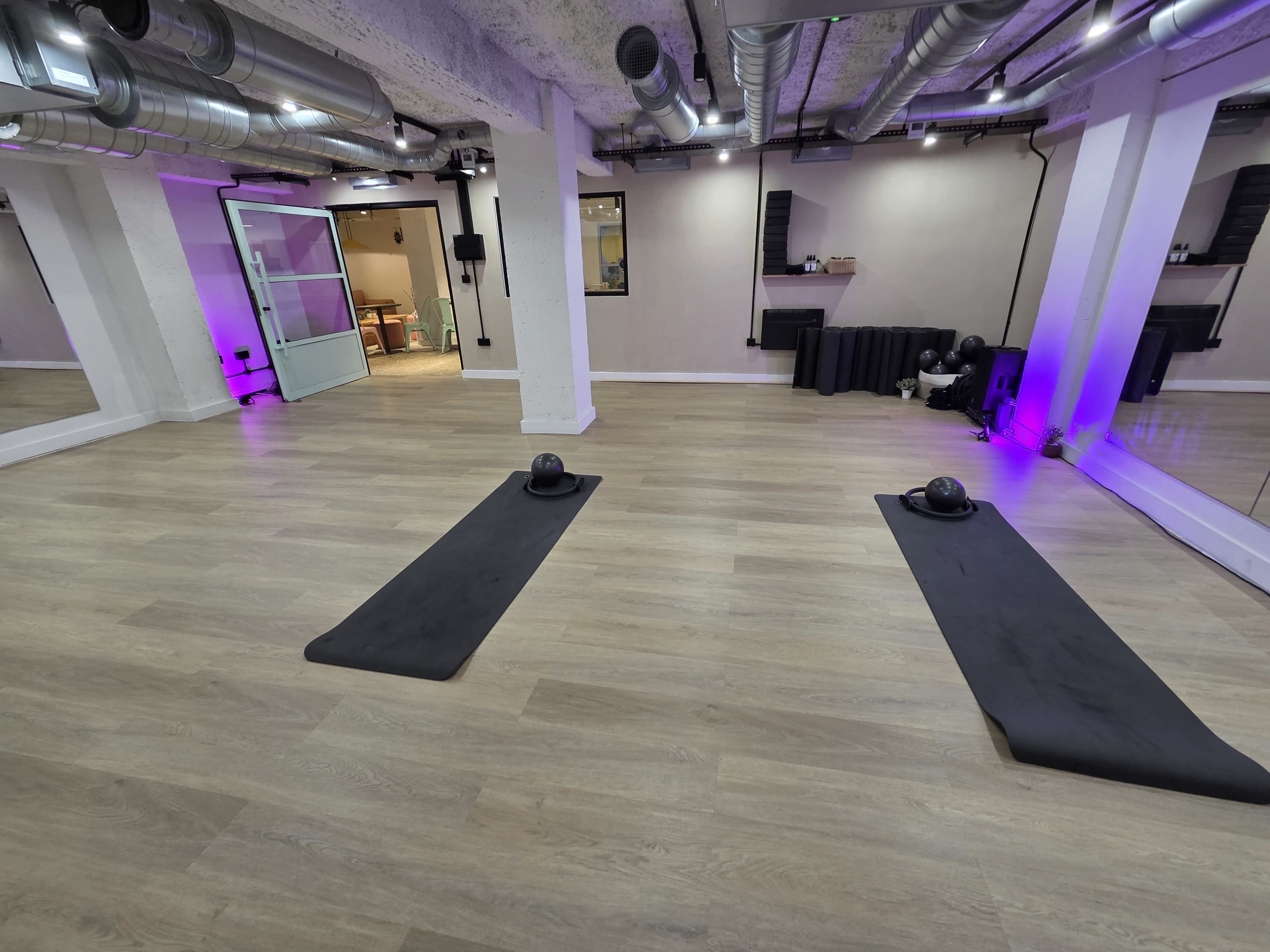 The image shows a spacious fitness studio with two exercise mats laid out on a wooden floor, dimly lit with purple lighting.