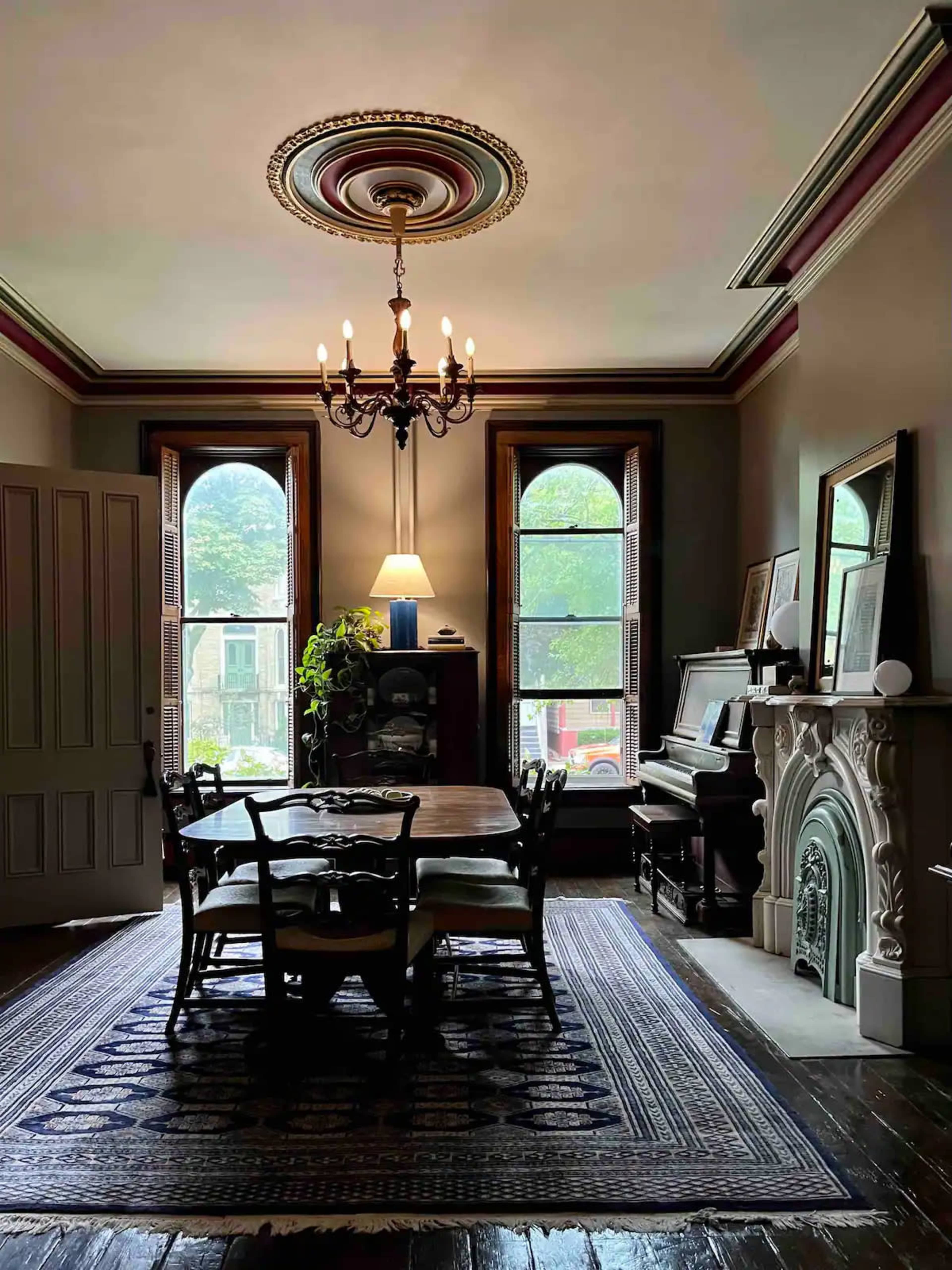 A dining room features a round table surrounded by chairs, with tall windows, a chandelier, and a decorative fireplace.