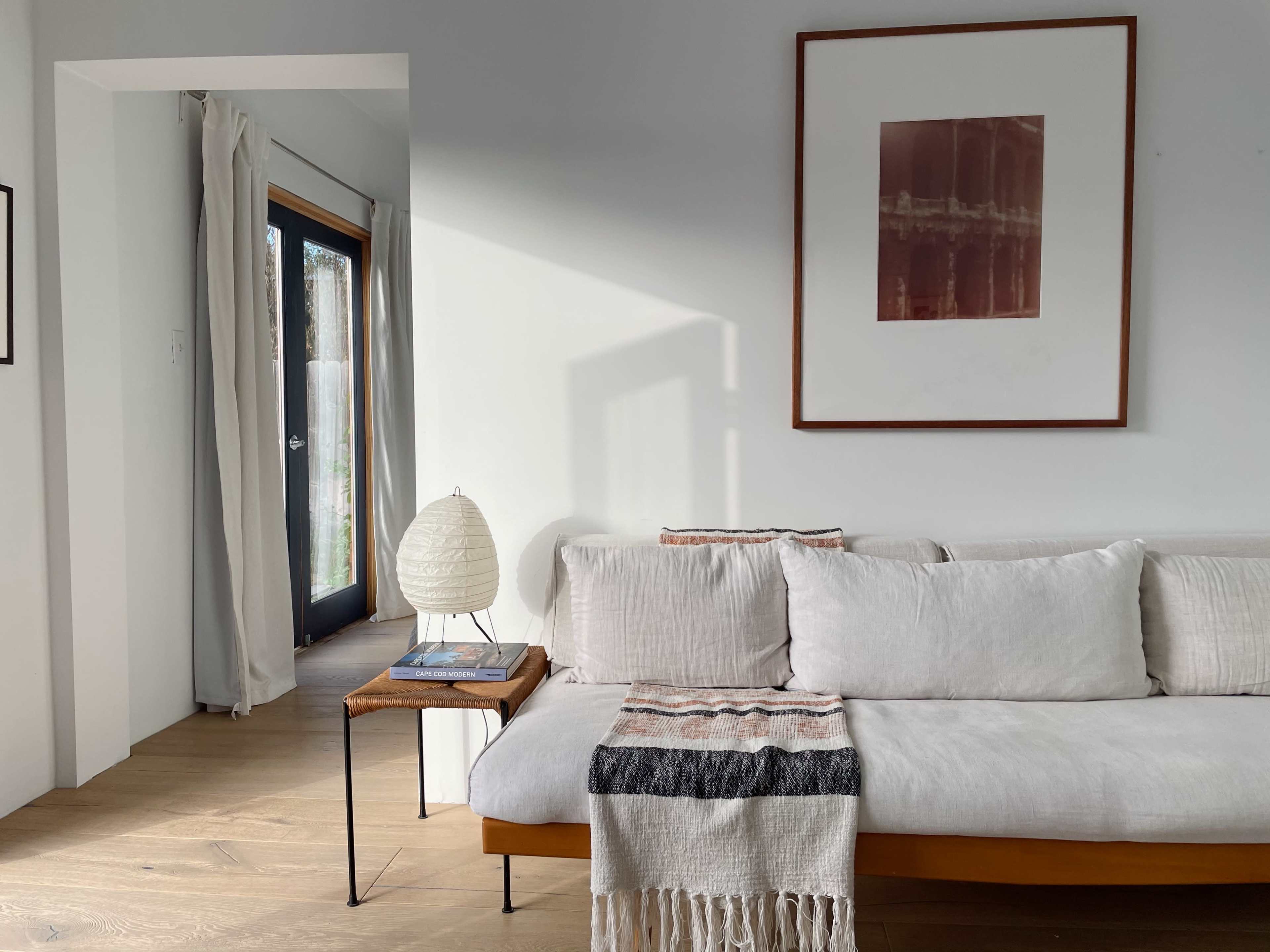 A light-filled living space features a sofa with neutral cushions, a small wooden side table, and a framed artwork on the wall.