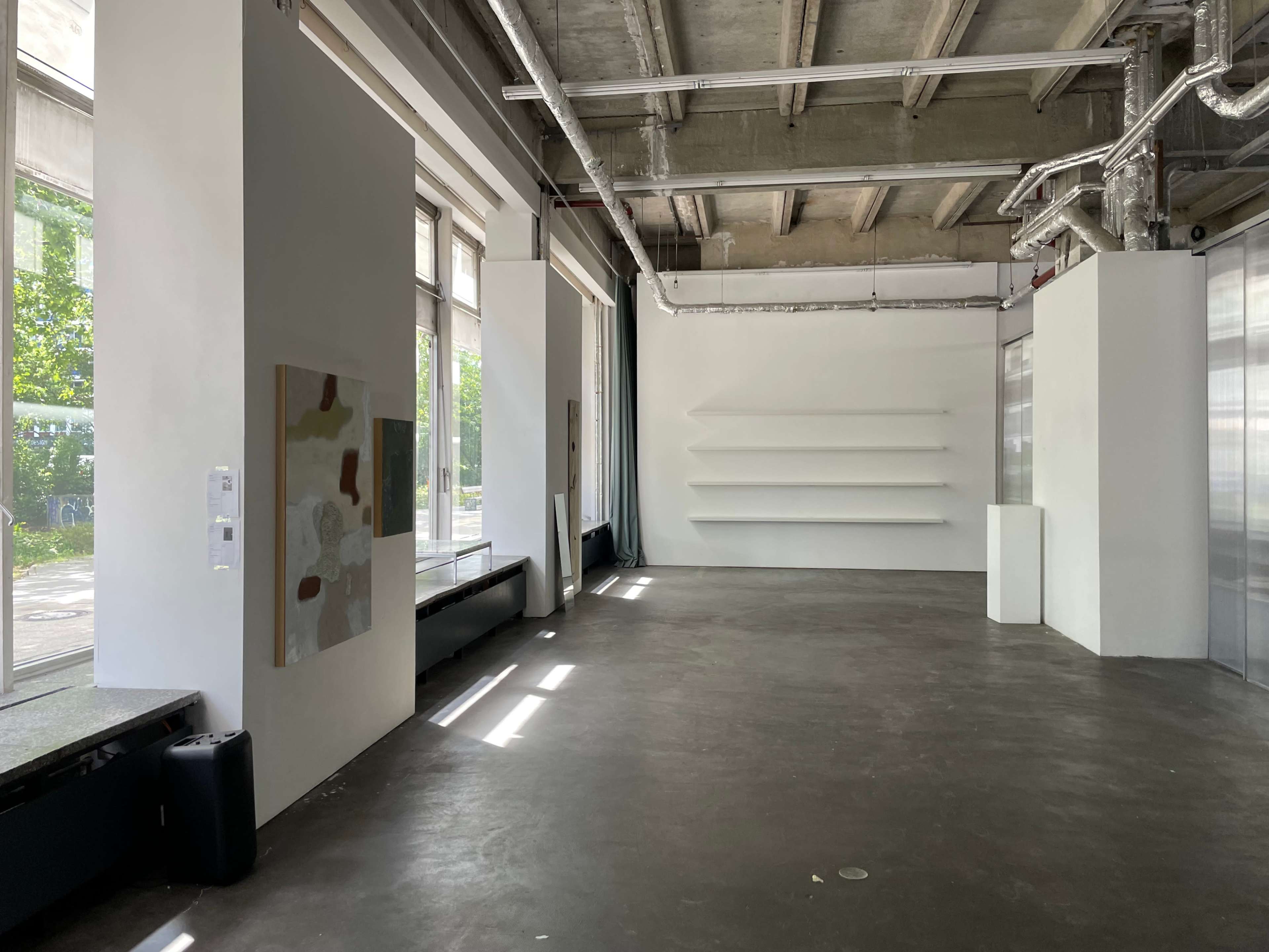The image shows a spacious, minimalist art gallery with large windows, concrete floors, and white walls, featuring a single artwork on display and empty shelving along one wall.