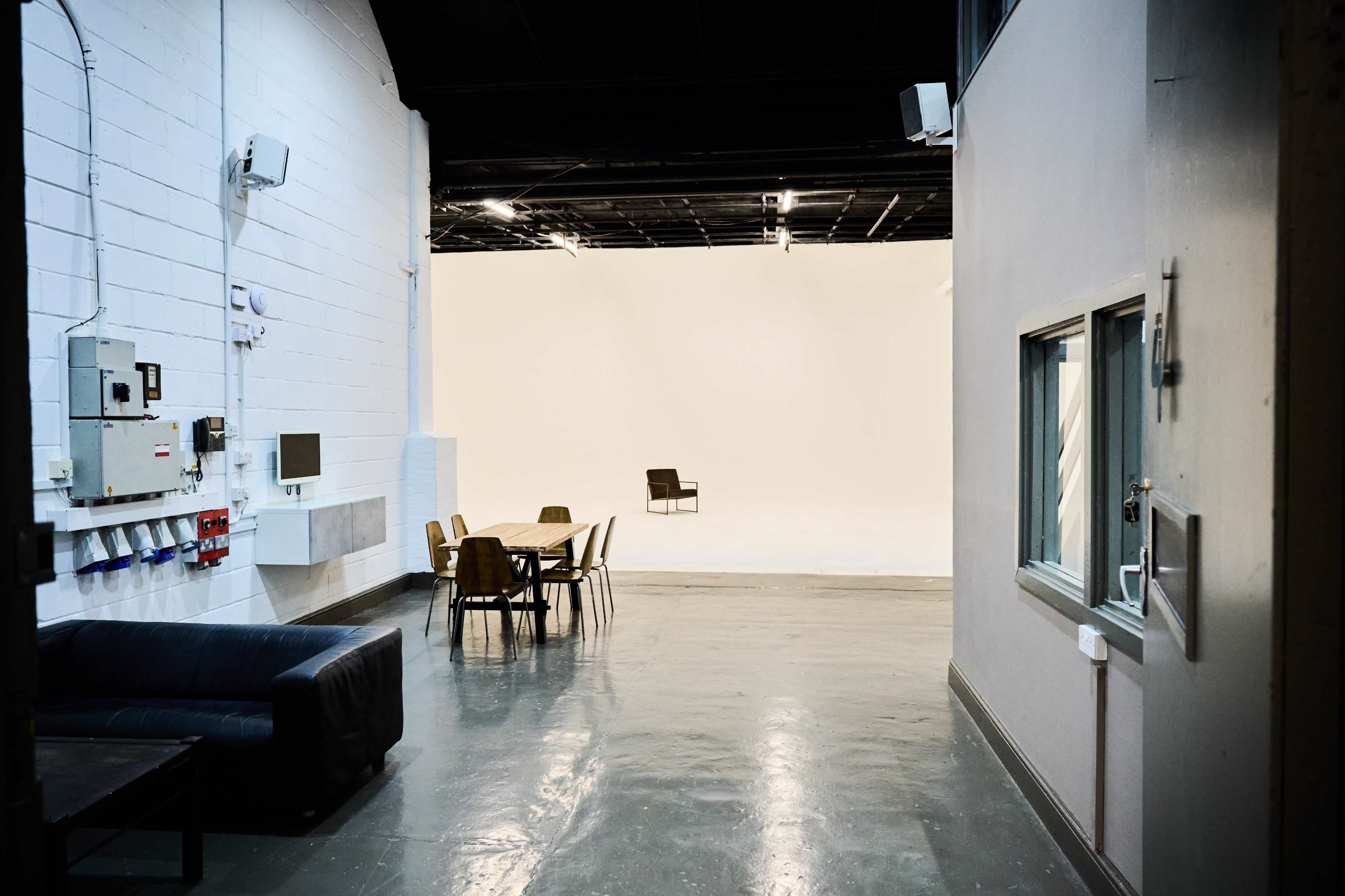 The image shows a spacious, well-lit studio with a plain white backdrop, a single chair in the center, and furniture including a table and chairs in the foreground.