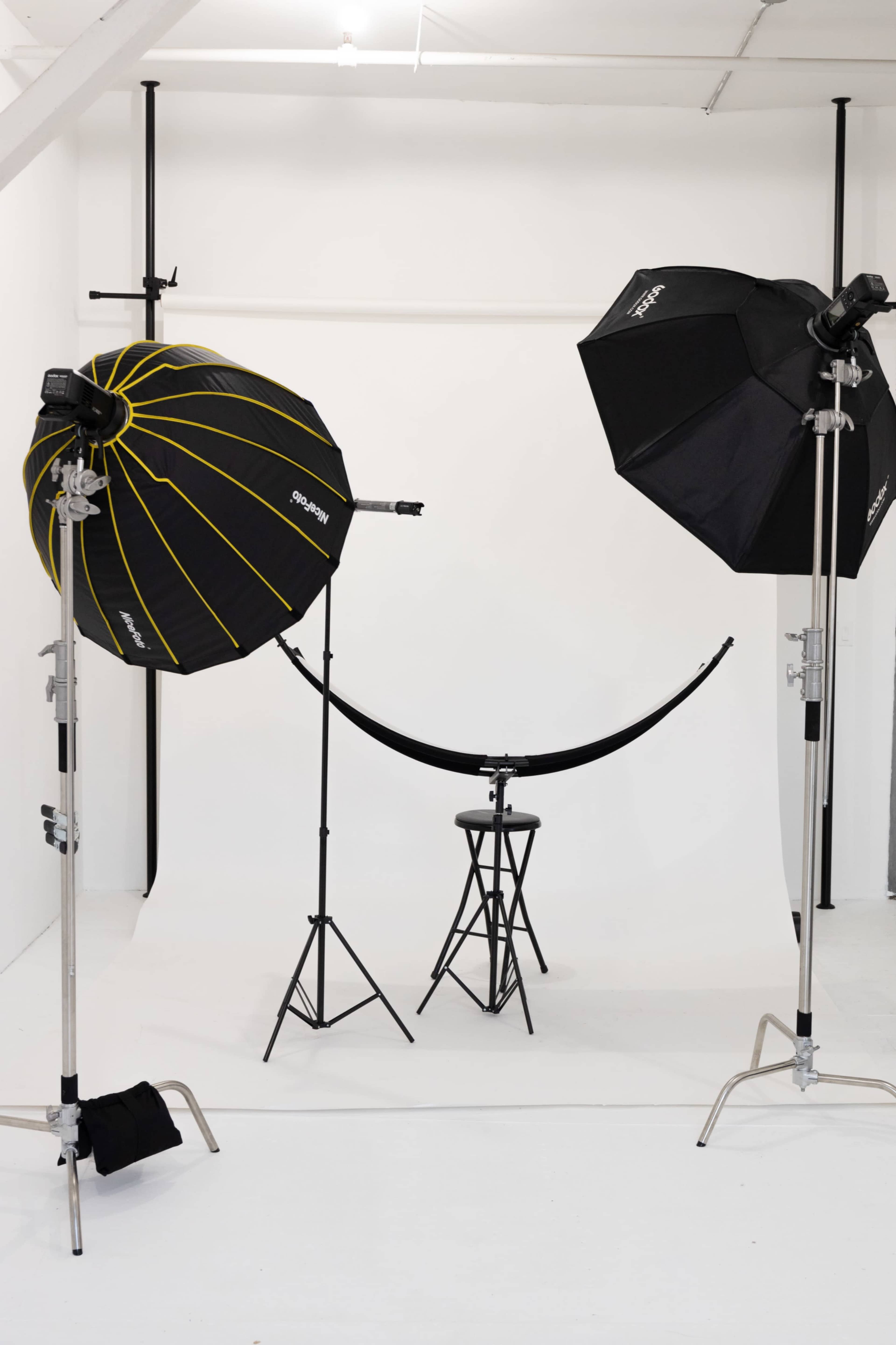The image shows a photography studio setup with two large softbox lights, a curved backdrop, and a small black stool in the center.