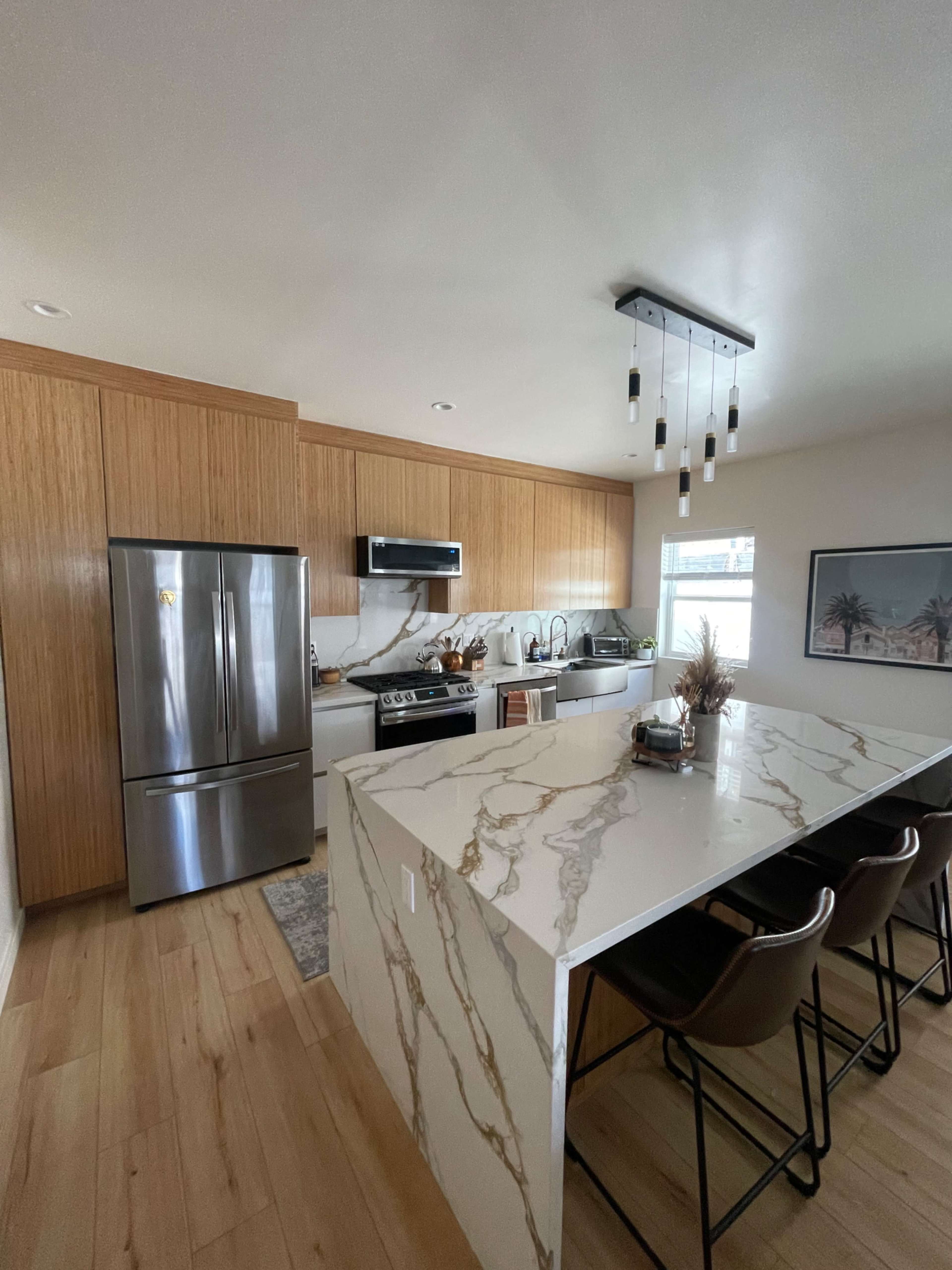 A modern kitchen features wooden cabinetry, stainless steel appliances, and a marble island with seating.