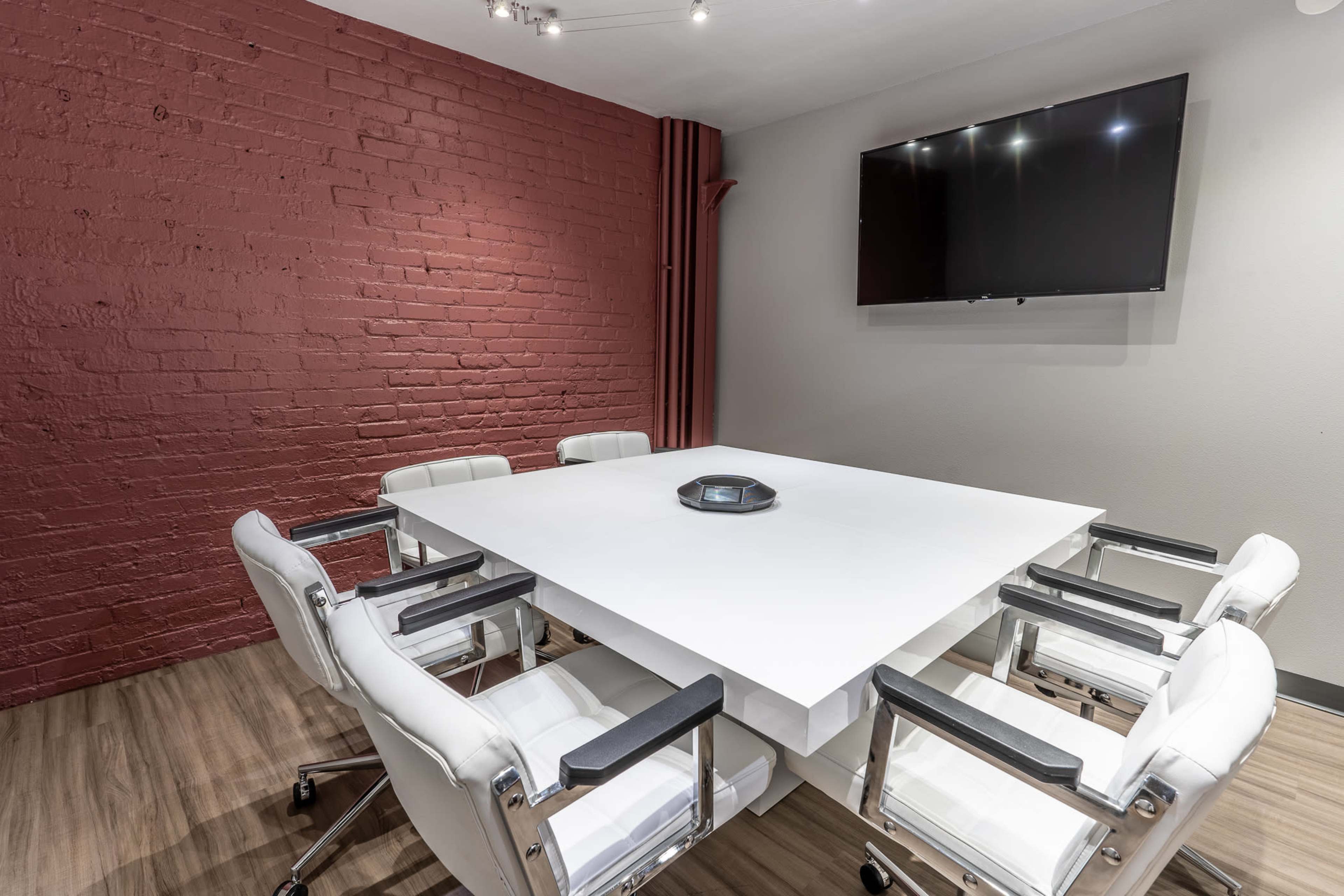 A modern conference room features a large white table surrounded by white chairs, with a red brick wall and a mounted TV.