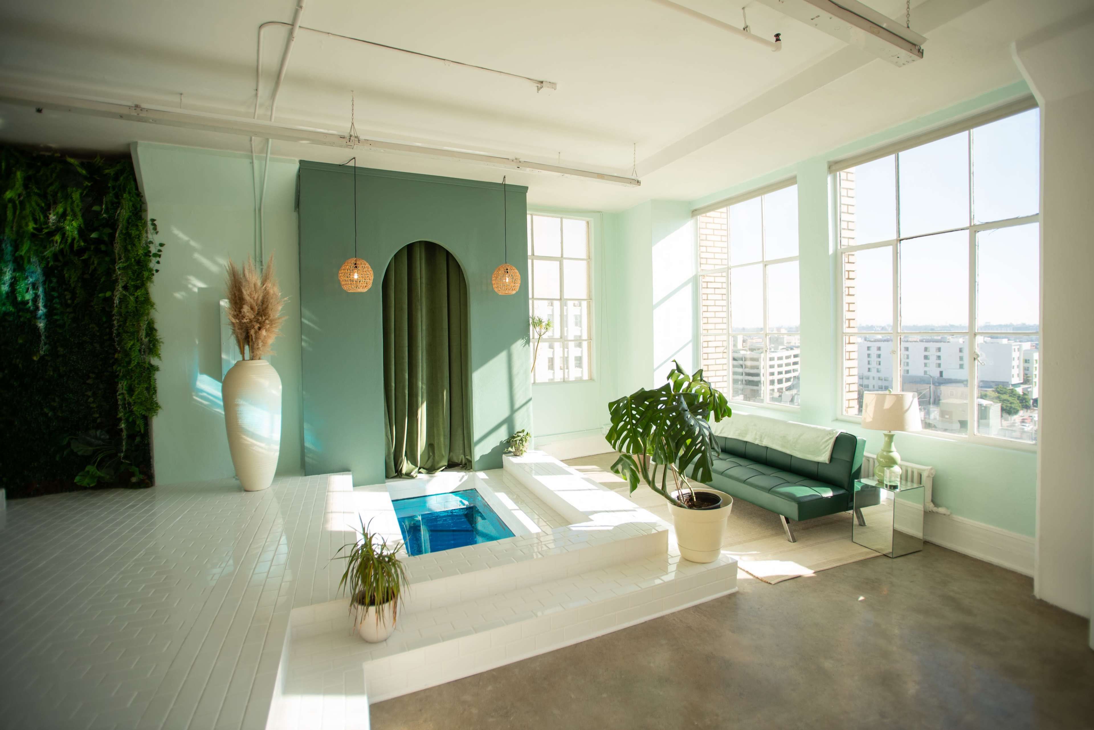 A modern interior space features a small indoor pool surrounded by plants and a turquoise wall, with large windows letting in natural light.