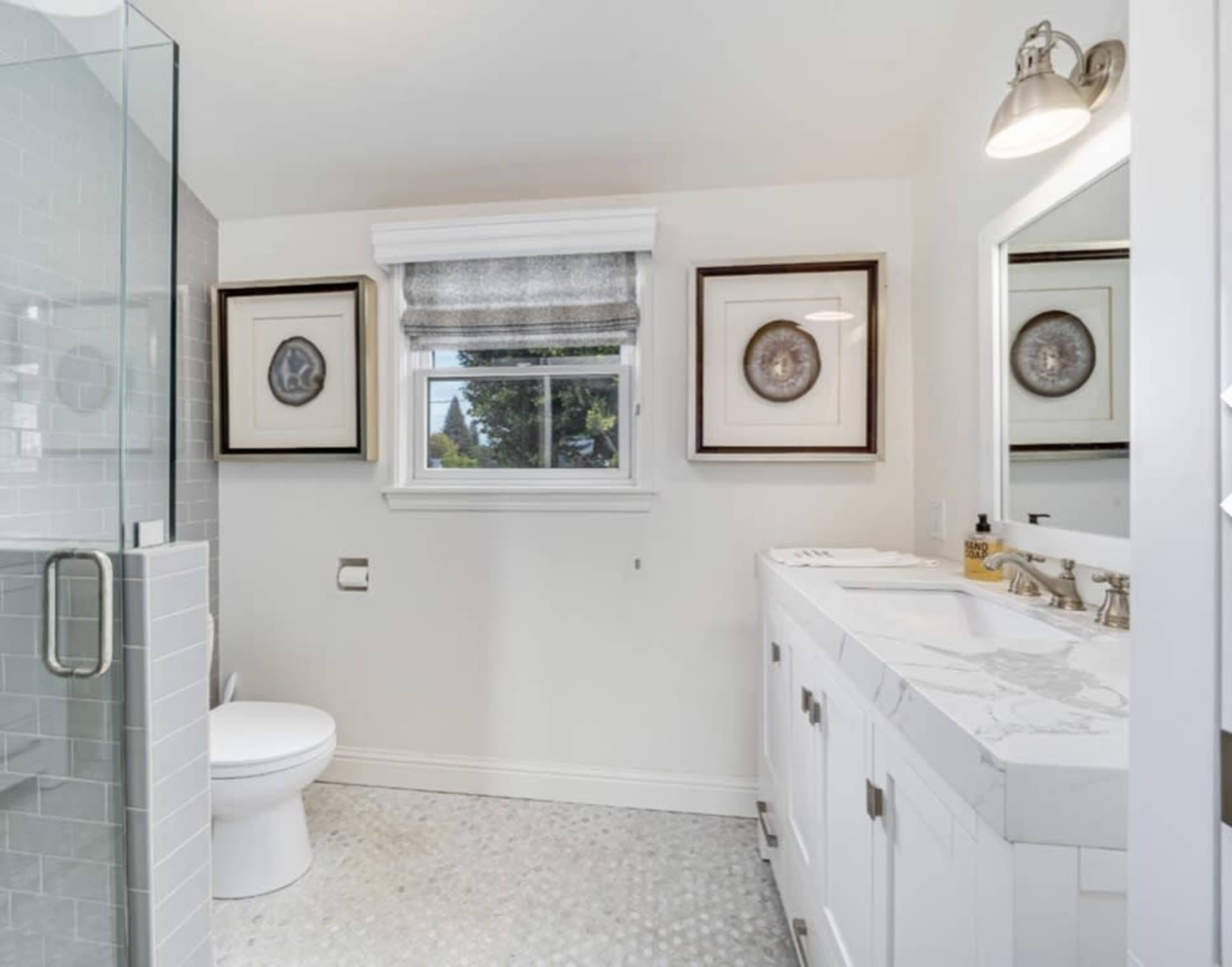 A modern bathroom featuring a glass shower, a white vanity with a marble countertop, and two framed circular artworks on the wall.