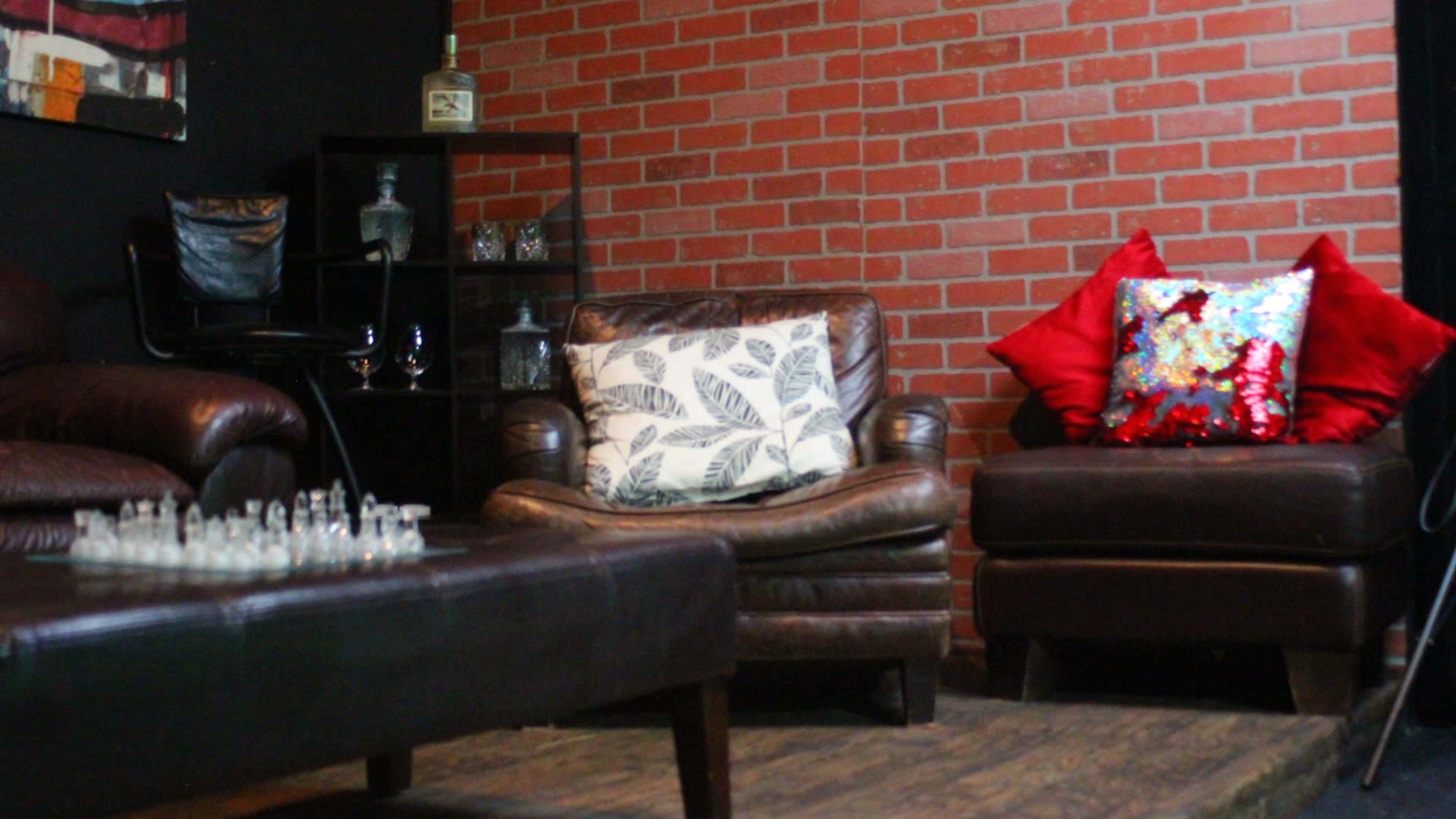 A cozy lounge area with leather chairs, a small table with a chessboard, and a shelf displaying glassware against a brick wall.