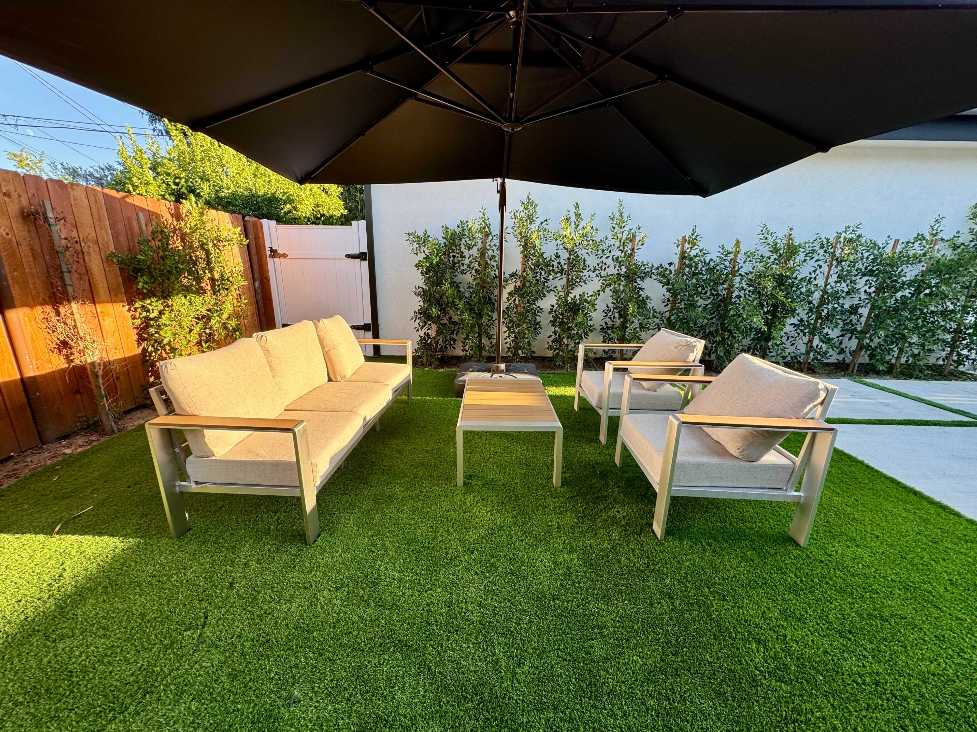A patio seating area features two sofas and two armchairs under a large umbrella, surrounded by green artificial grass and a fence lined with shrubbery.