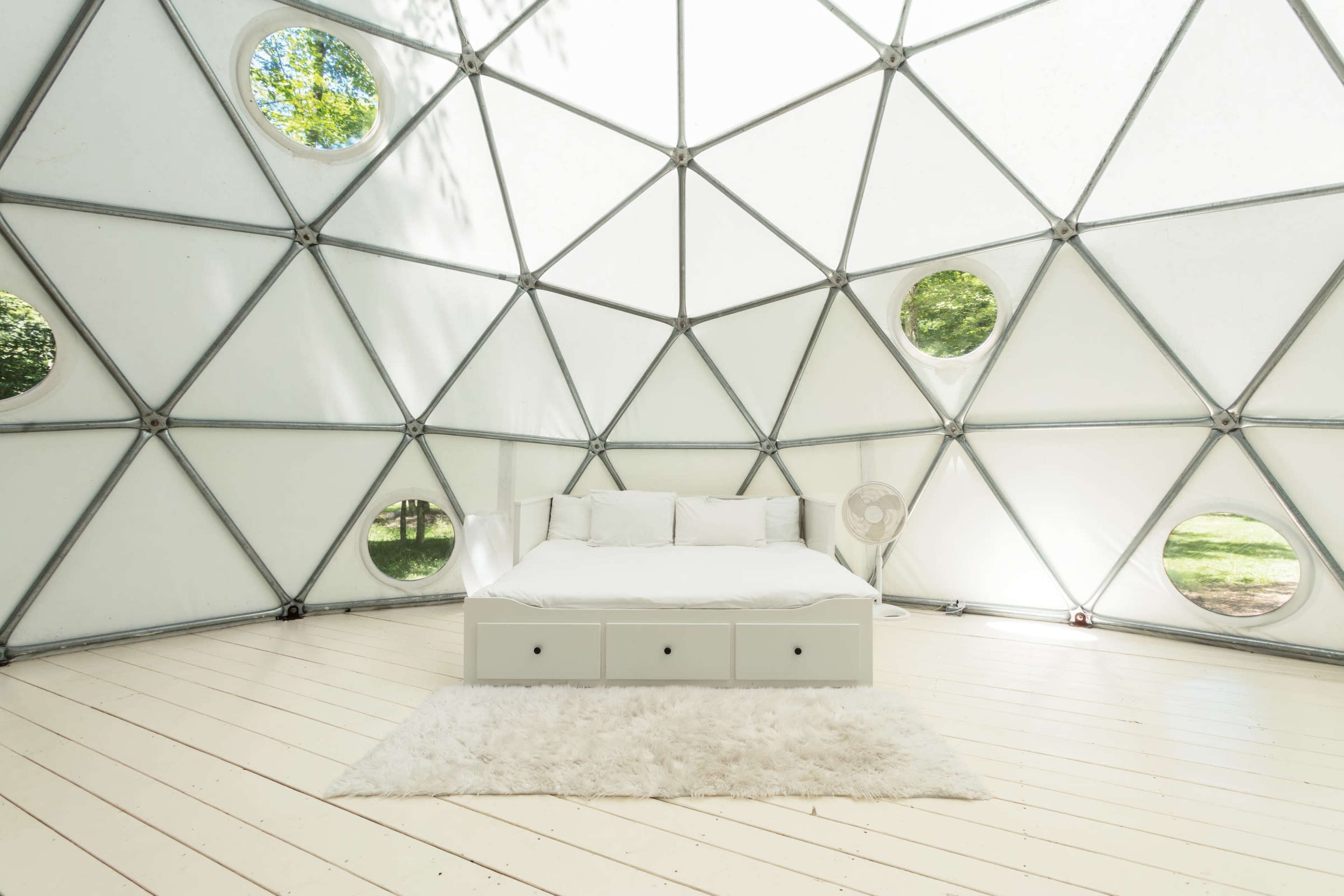 The image shows a minimalist geodesic dome interior featuring a bed with a white cover and several round windows allowing natural light to enter.