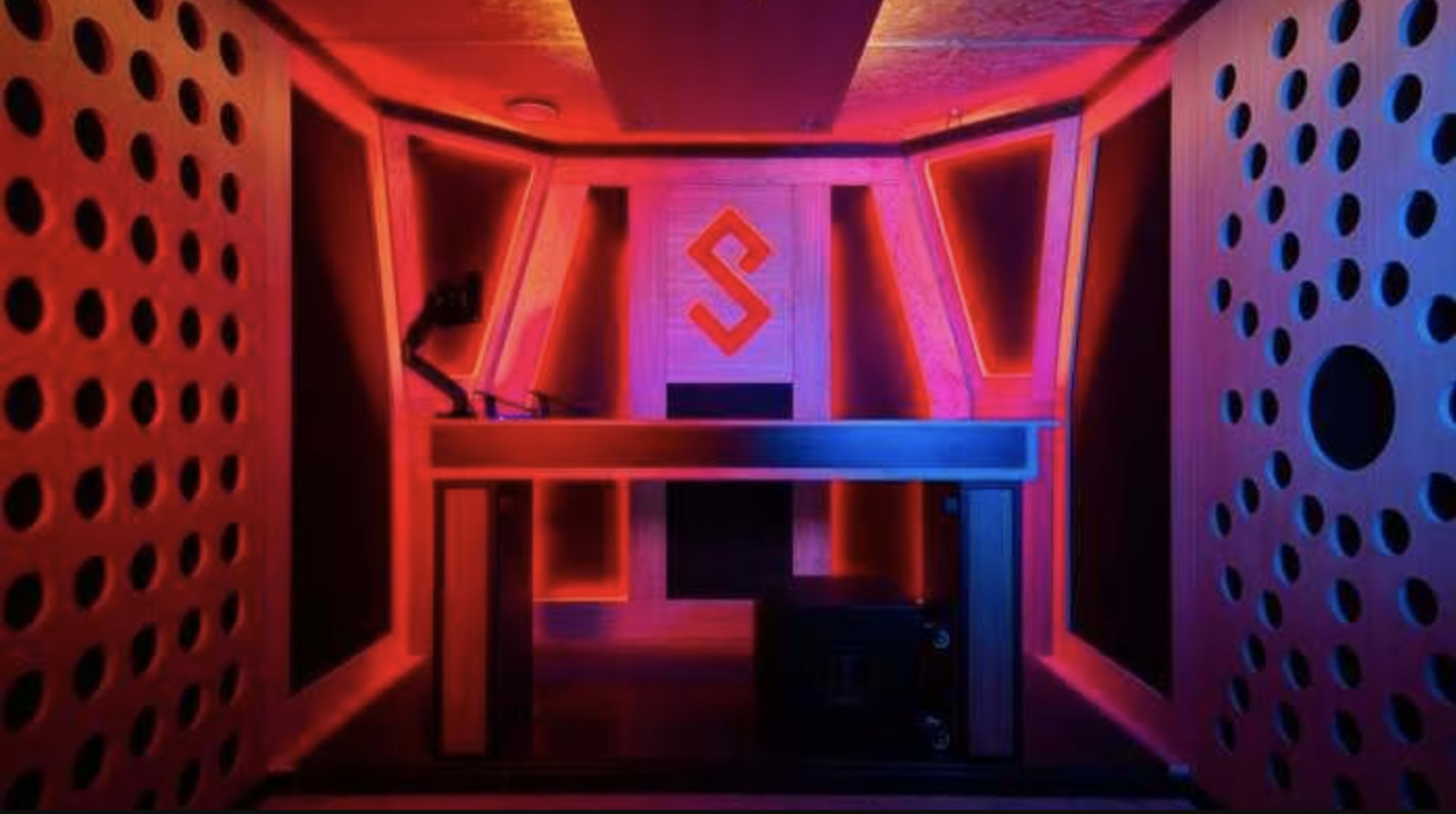 A room with a futuristic design featuring orange and blue lighting, a raised platform with equipment, and patterned walls.