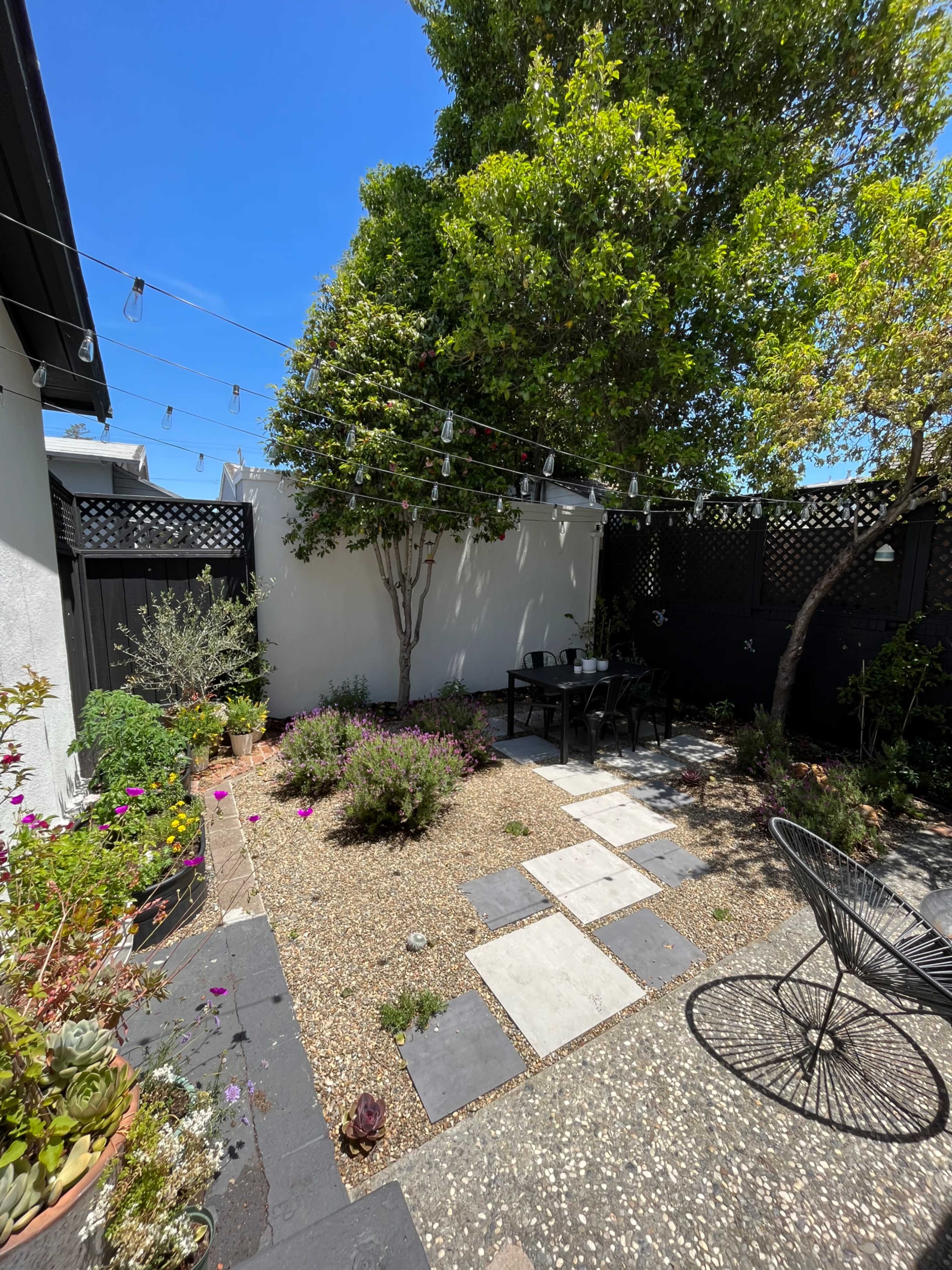 A small garden features gravel paths, a stone table with chairs, and various plants under a clear blue sky.