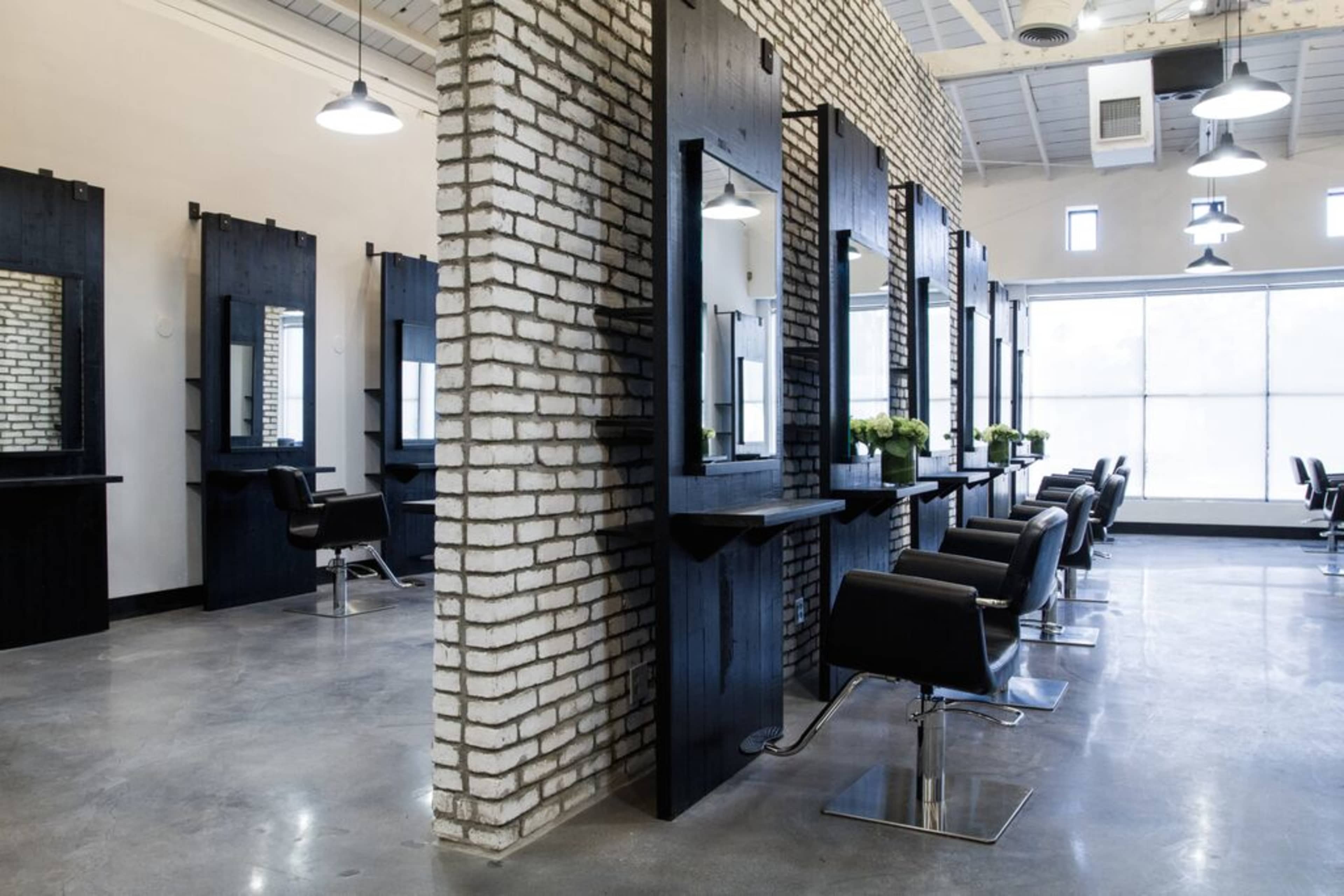 The image shows a modern hair salon featuring black wooden styling stations with mirrors, sleek black chairs, and a brick wall, set in a spacious, well-lit environment.