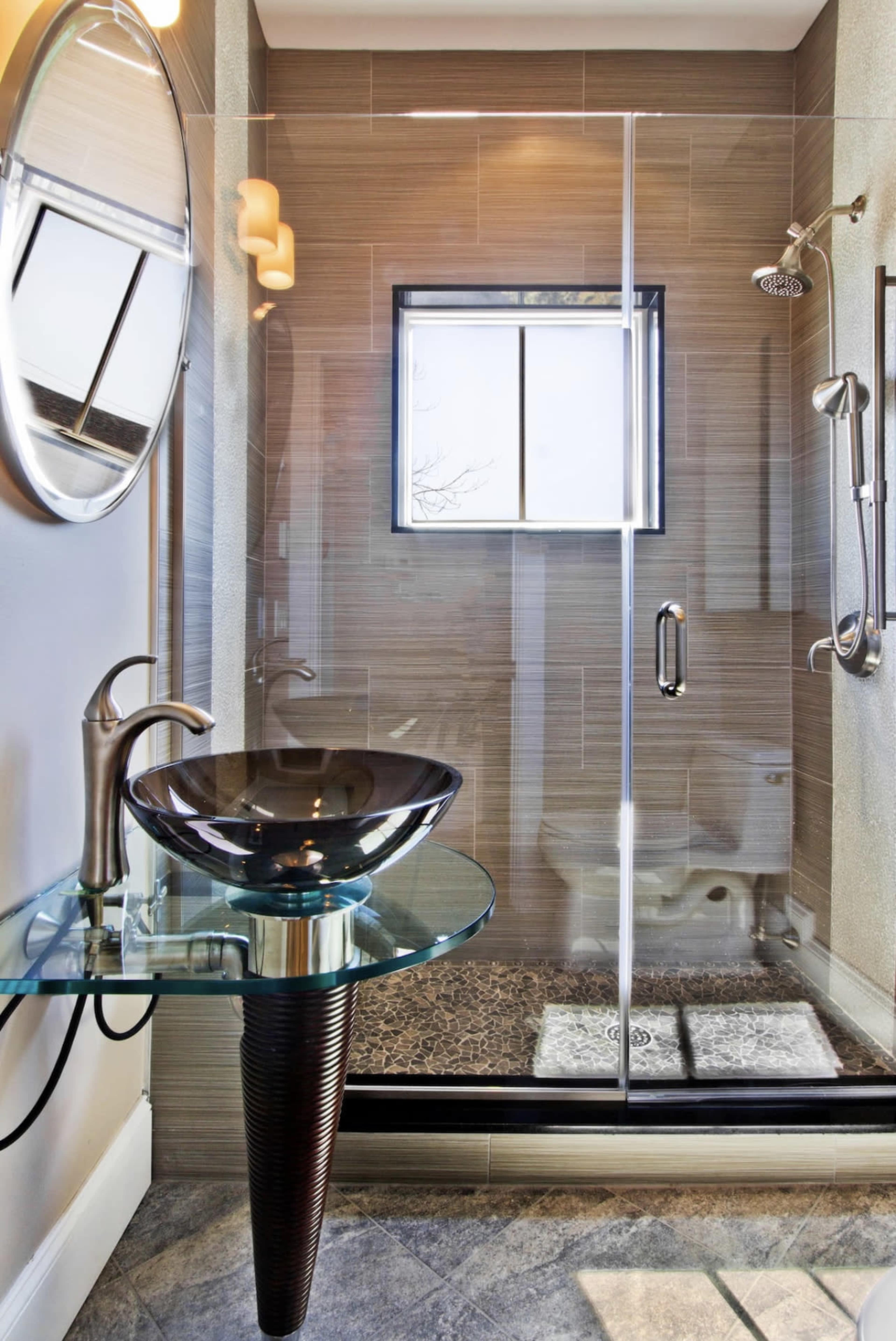 A modern bathroom features a glass-enclosed shower, a round mirror, and a sleek, black vessel sink on a glass countertop.