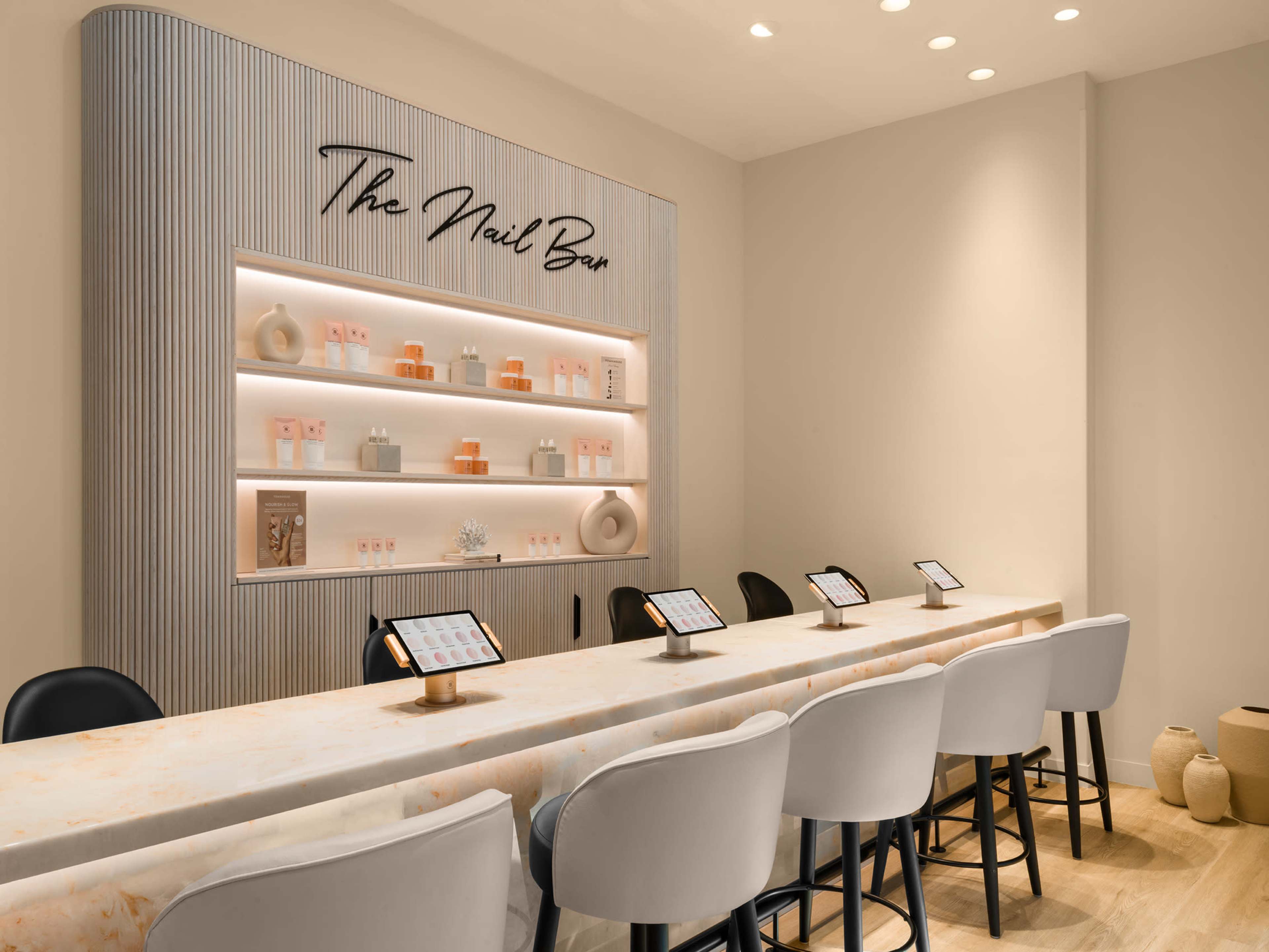 The image shows a modern nail bar with a long, marble-topped table, equipped with several tablets for customer use, and a neatly arranged shelf displaying various beauty products.