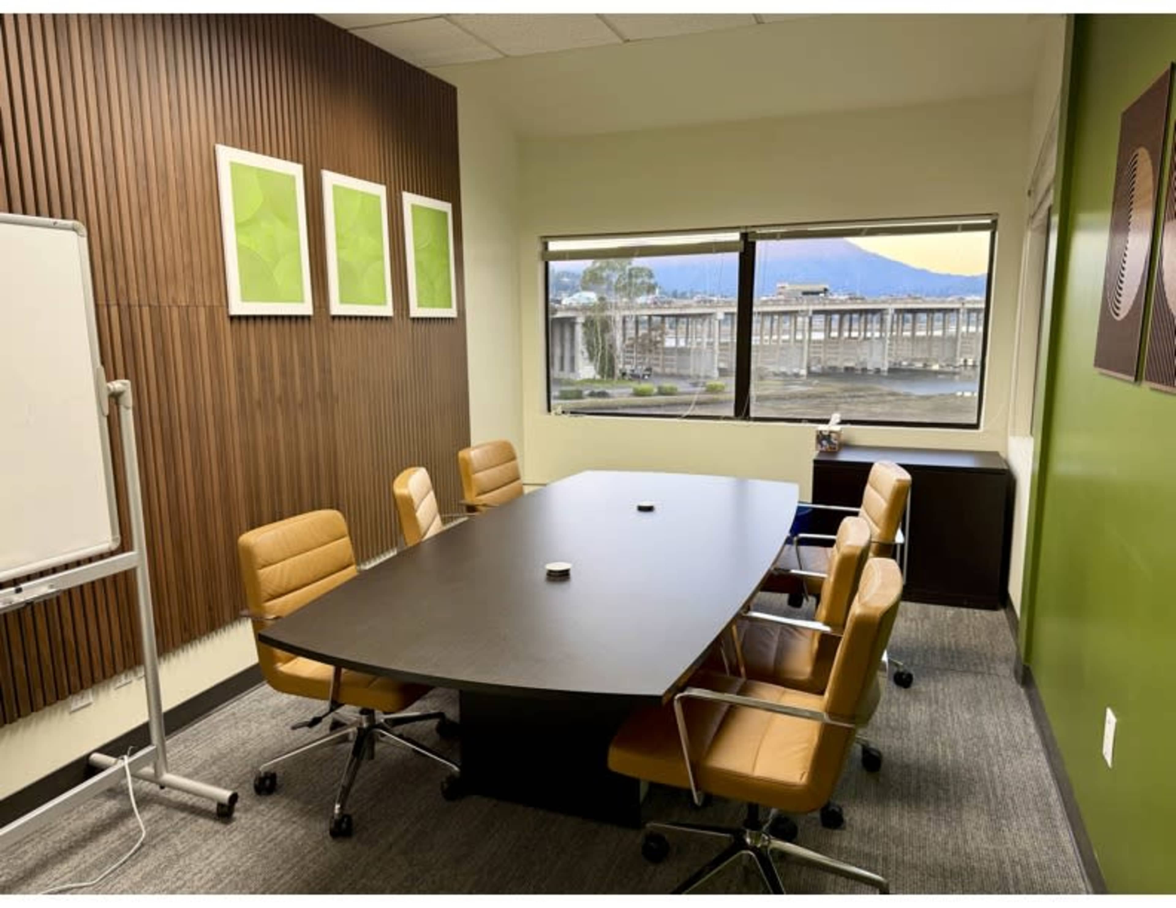 The image shows a conference room with a large oval table surrounded by four leather chairs, a whiteboard, and a window displaying a view of a river and bridge.