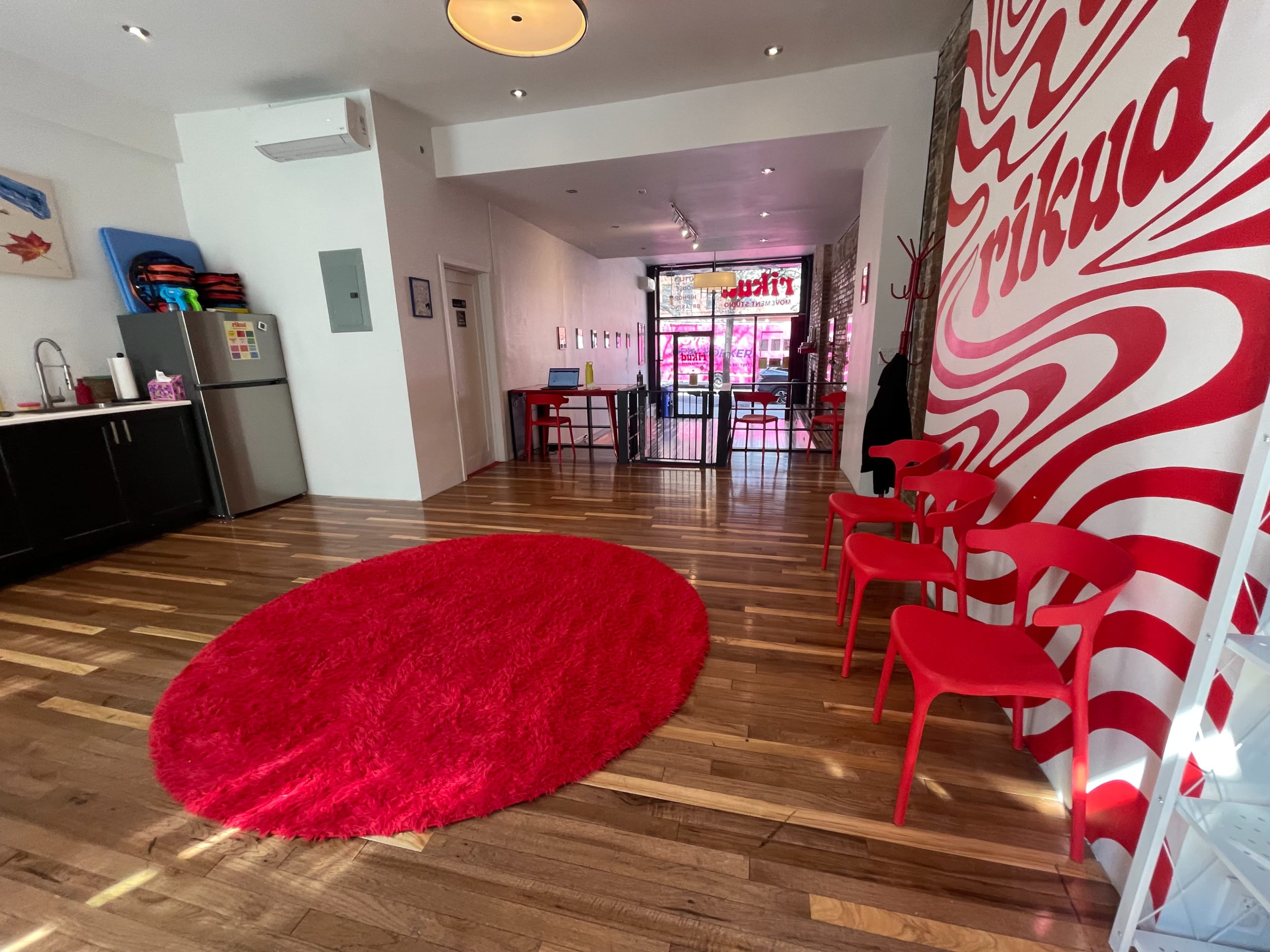 The image shows a modern interior space with a red circular rug and red chairs, featuring a distinctive white and red patterned wall and visible entrance to the street.
