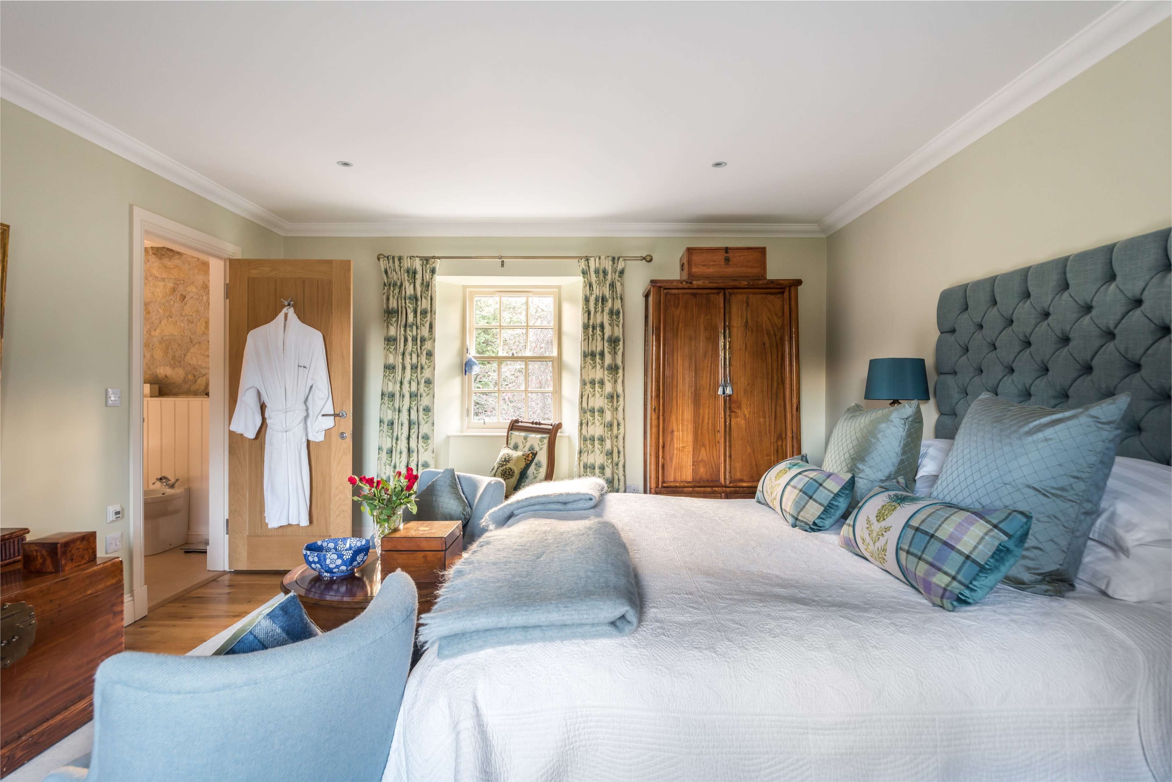 A well-furnished bedroom featuring a large bed with decorative pillows, a wooden wardrobe, and a cozy seating area, illuminated by natural light from a window.