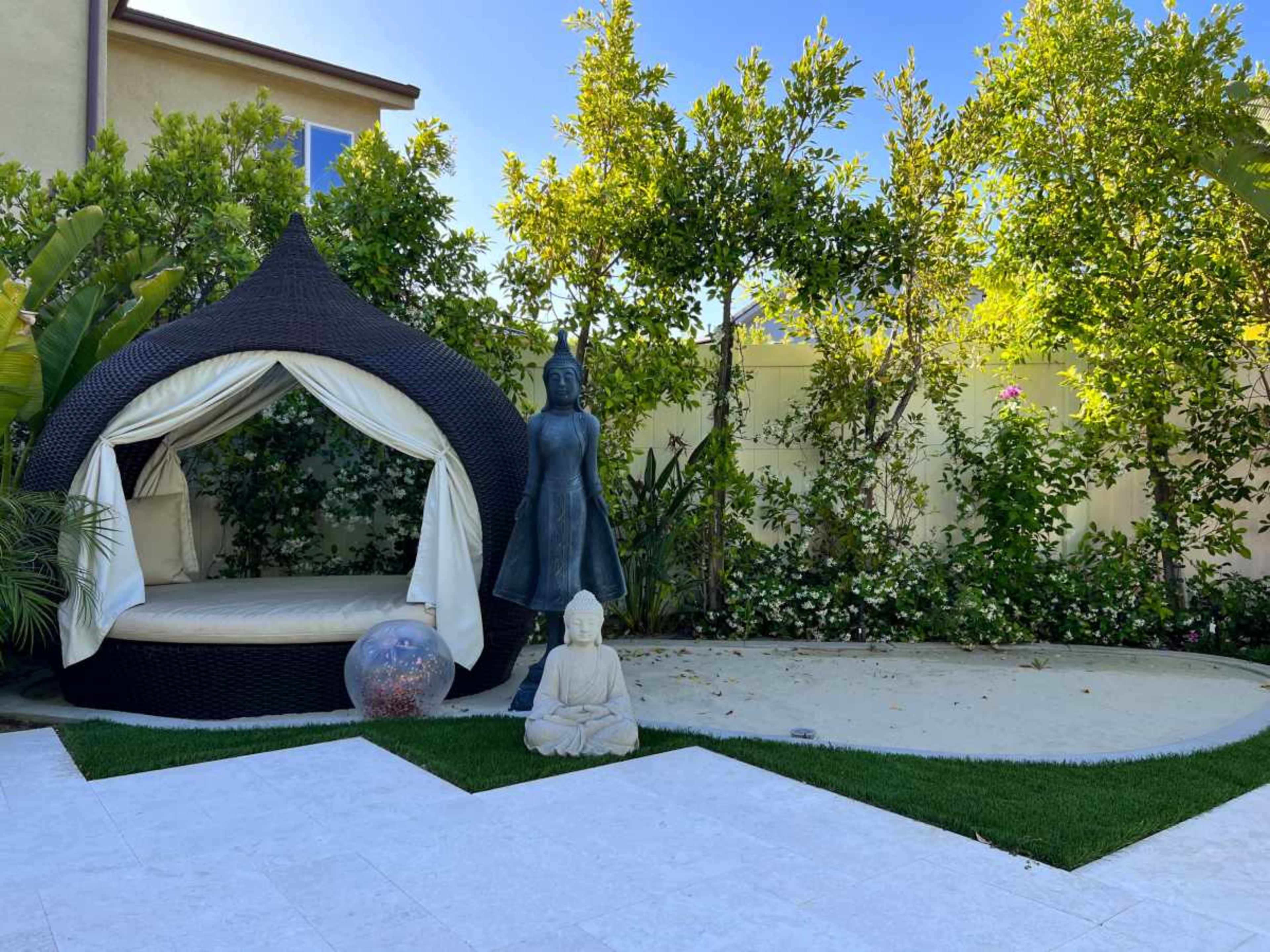 The scene features a shaded lounge area with a curved black wicker structure, surrounded by greenery and decorative statues in a landscaped yard.