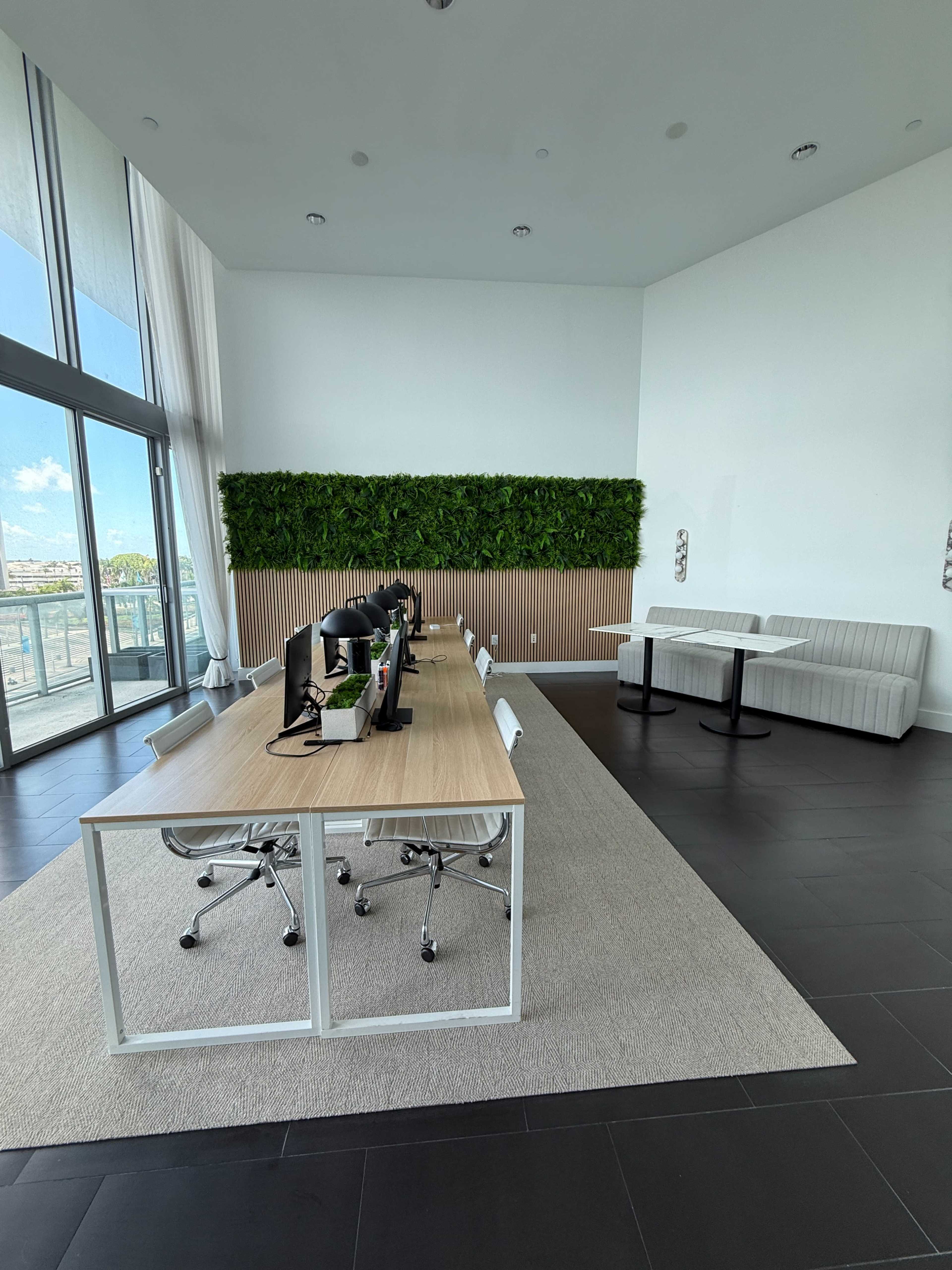 The image shows a modern office space featuring a long wooden table with several computer monitors, a green plant wall, and a seating area with two black tables and a light-colored couch.