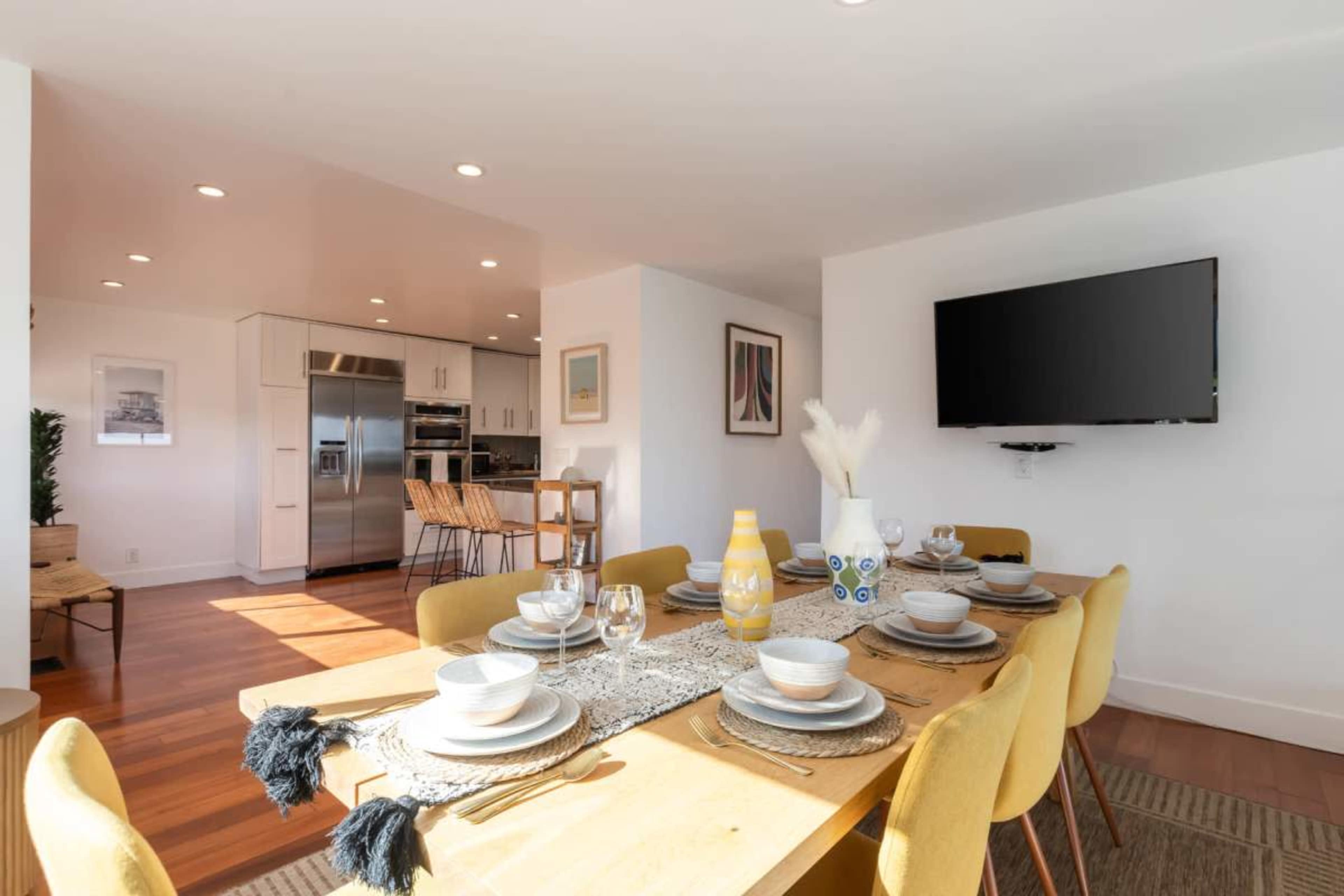 A dining area features a large wooden table set with dishes and utensils, adjacent to a modern kitchen with stainless steel appliances and a wall-mounted television.