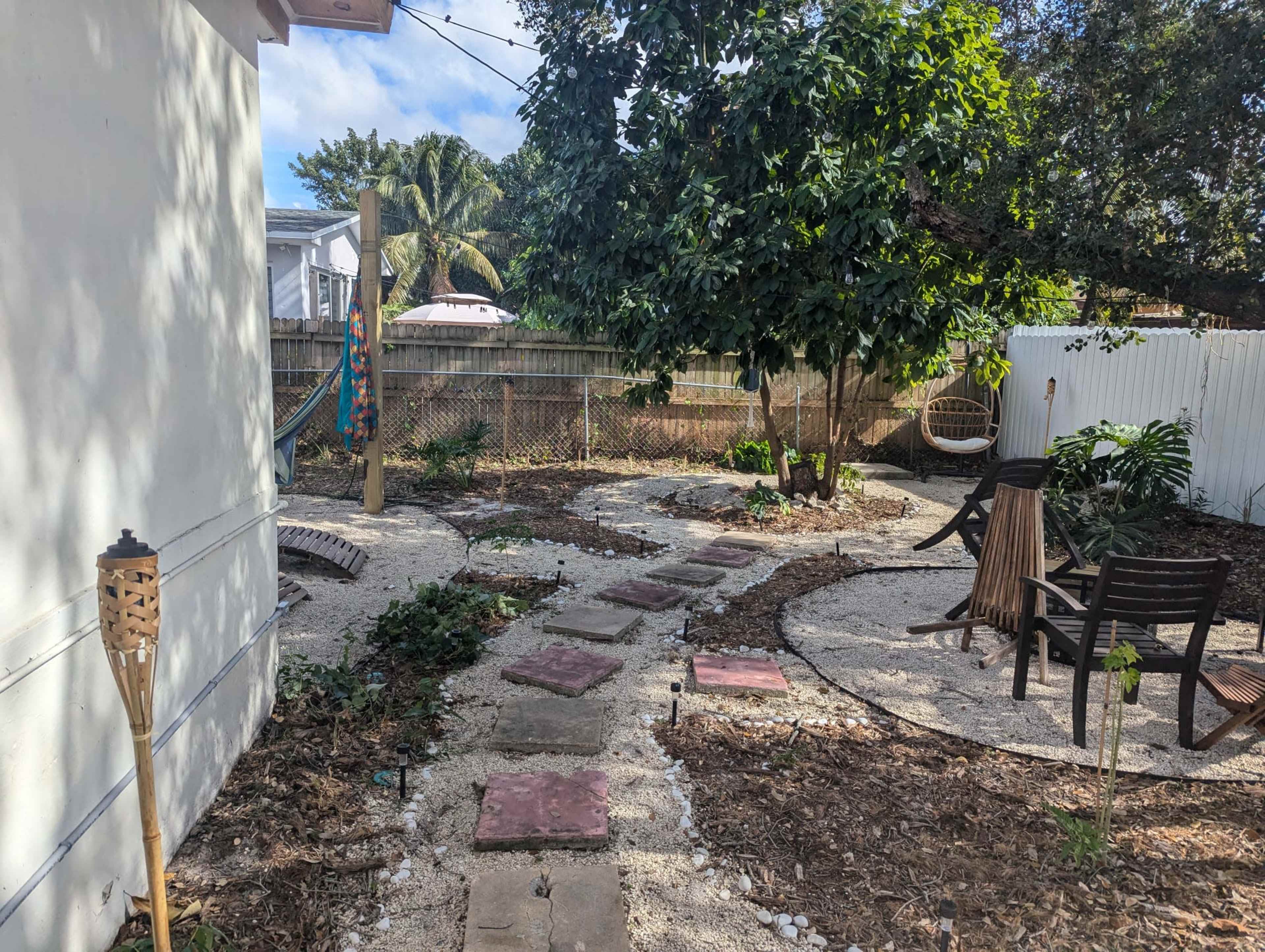 The image shows a landscaped garden area with a winding stone path, surrounded by plants, a hammock, and seating arrangements under a large tree.