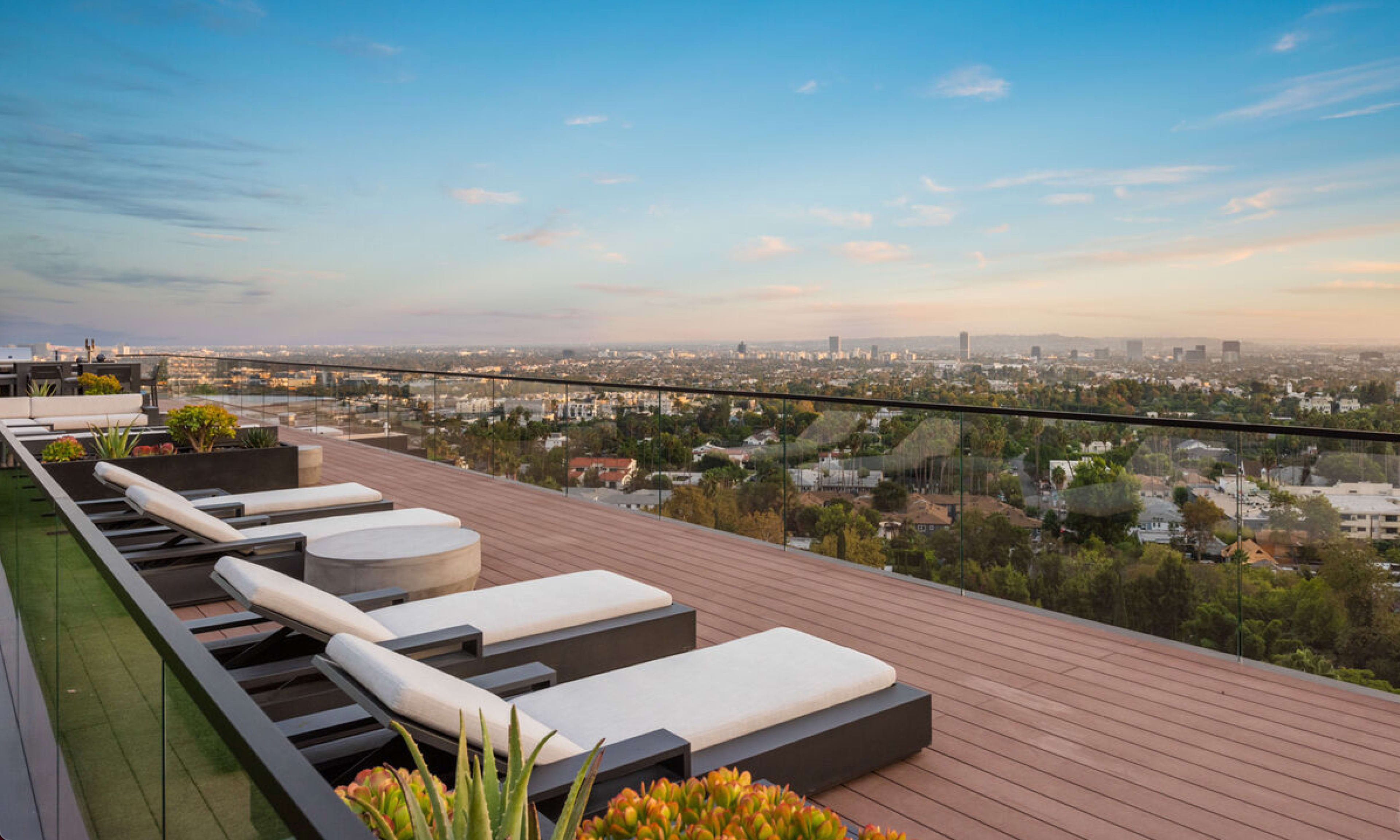 The image shows a modern balcony with lounge chairs overlooking a city skyline at sunset.