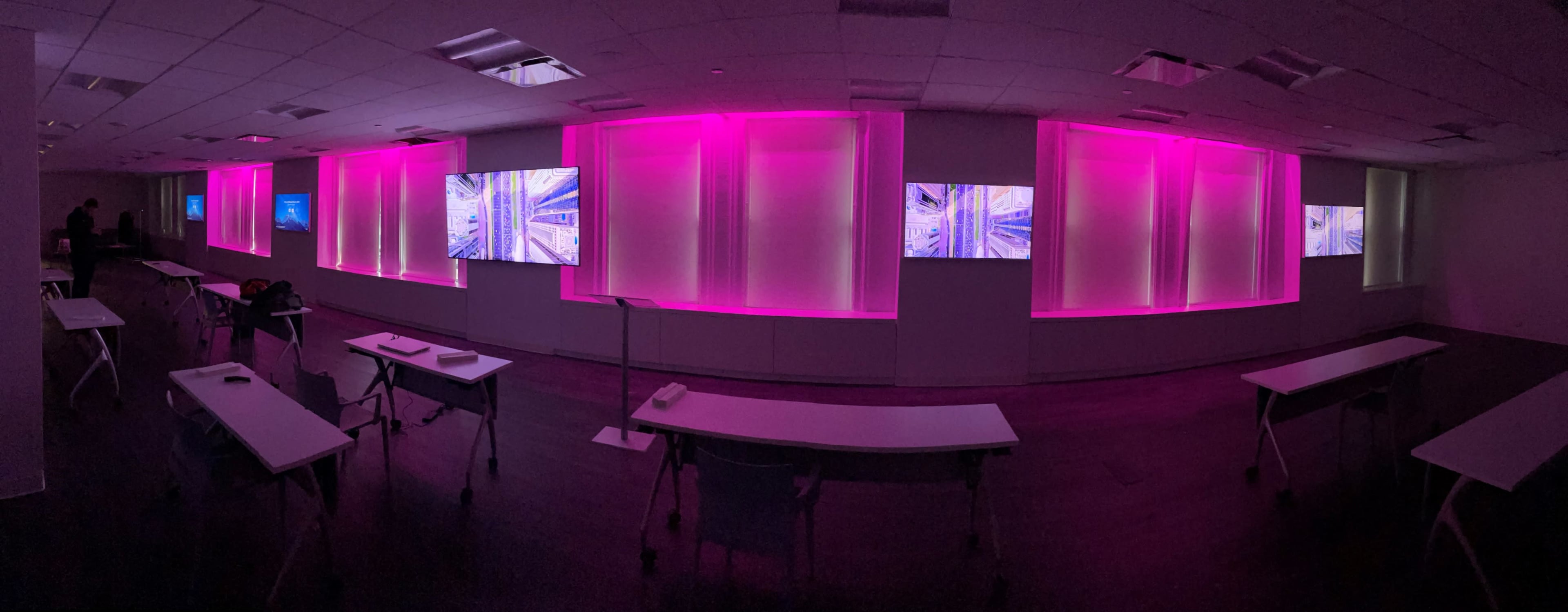 A modern, dimly lit room with multiple screens displaying abstract visuals, surrounded by pink ambient lighting and arranged tables facing the displays.