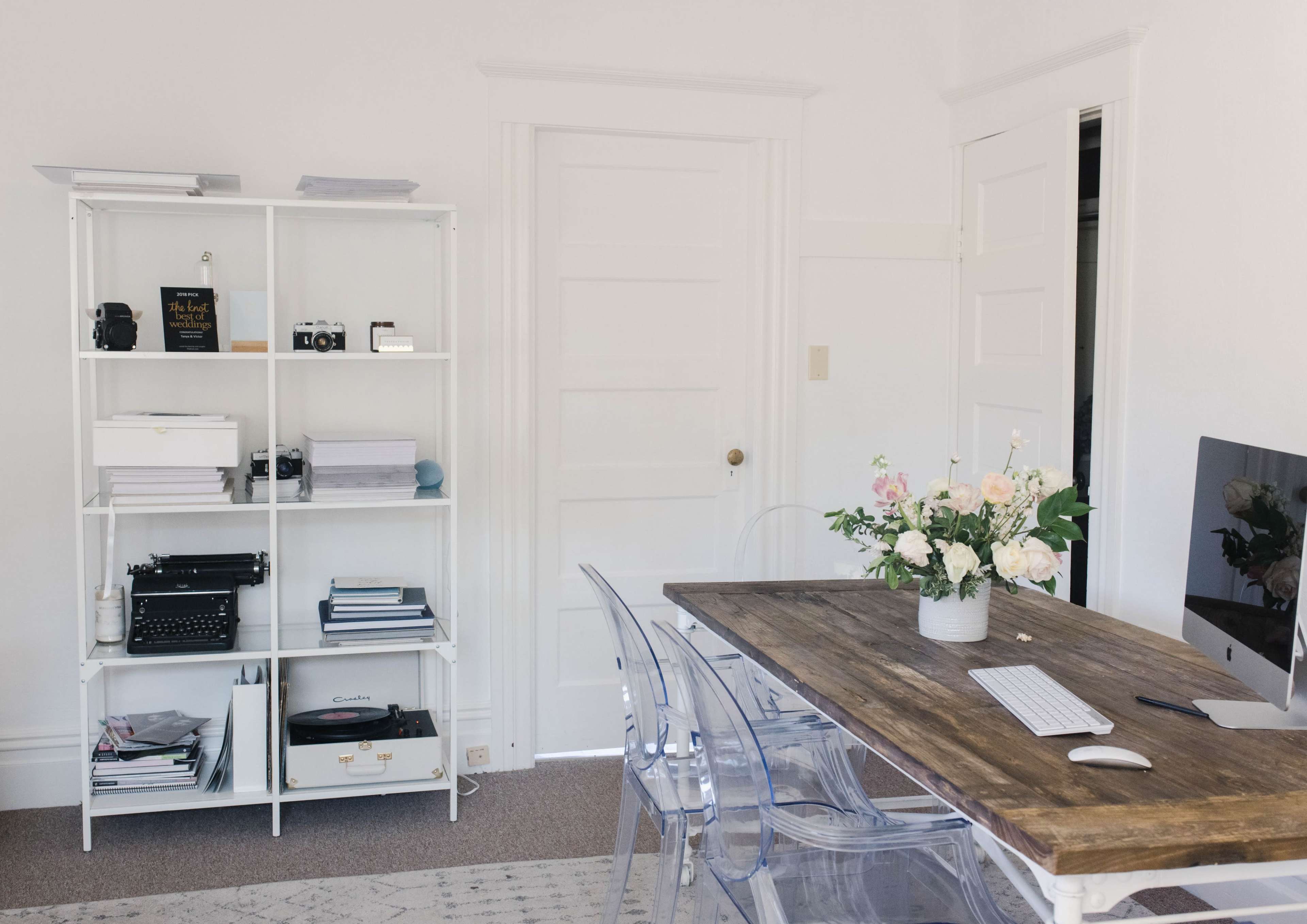 A minimalist workspace features a wooden table, an iMac, a clear acrylic chair, and a white shelf filled with cameras and office supplies, along with a floral arrangement.