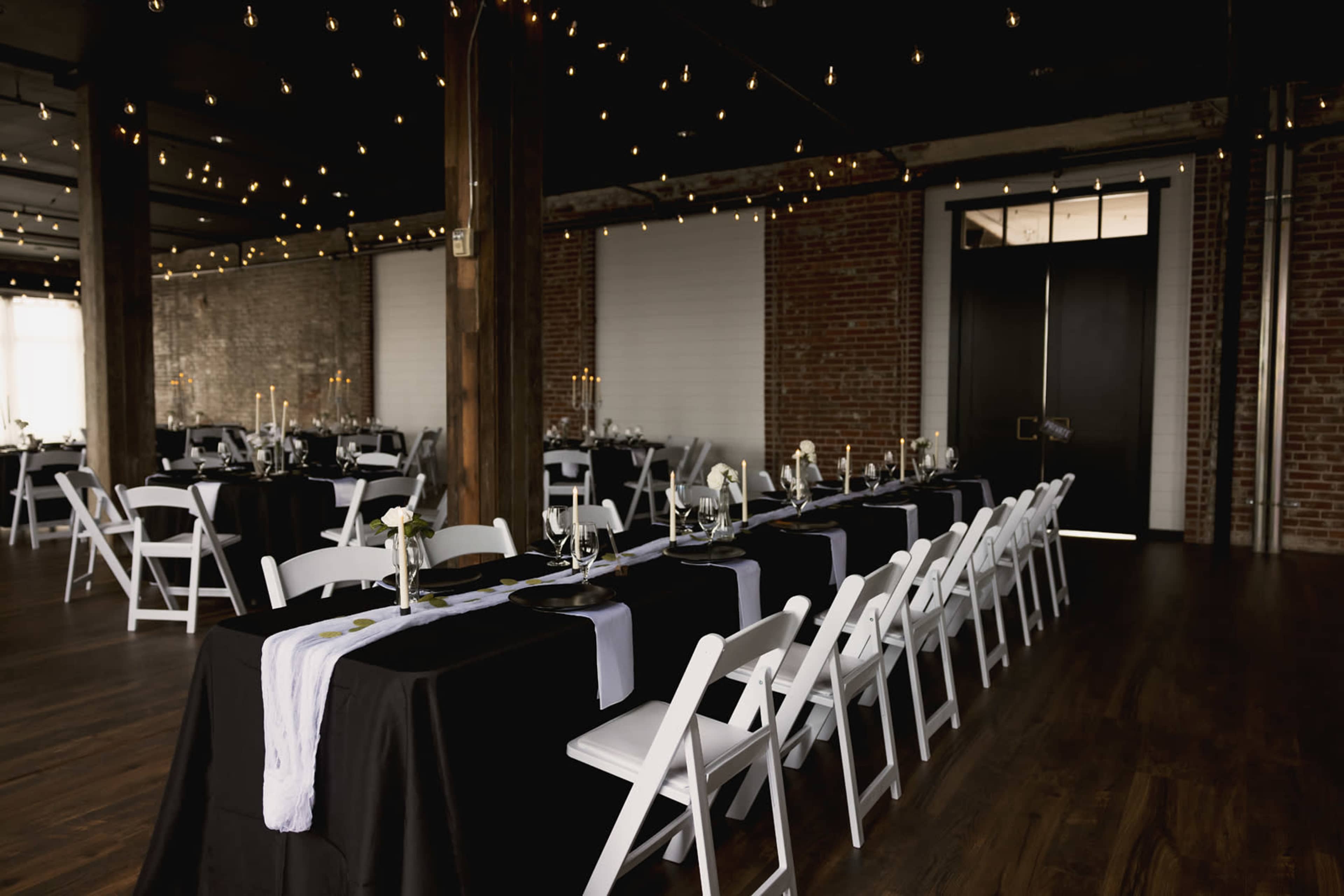 A decorated event space with long tables set with black and white tablecloths, white chairs, and ambient lighting from hanging string lights.